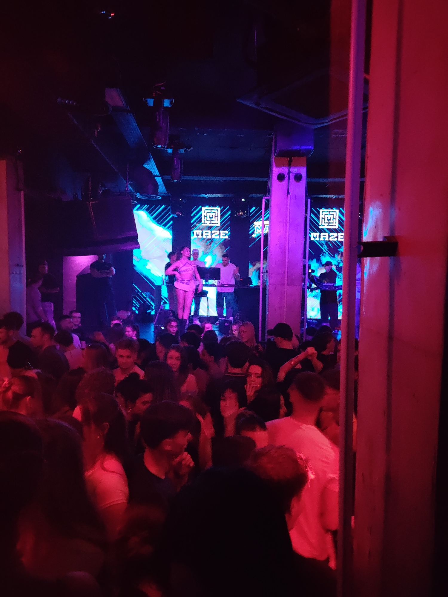 The image depicts the interior of Maze Club, a nightclub located in Varna, Bulgaria. The scene is densely populated with an estimated one hundred individuals, predominantly young adults, gathered in a dimly lit, high-ceilinged space. The primary illumination consists of strong magenta, purple, and blue stage lighting, casting deep red hues on the right foreground and vibrant purples on the stage area.
In the background, a raised stage features multiple vertical LED screens displaying the "MAZE" logo alongside dynamic blue and purple abstract patterns. On the stage, a female individual, dressed in a light-colored two-piece outfit resembling a cheerleading uniform with the number "22" visible on the top, stands centrally, facing the crowd. To her left (viewer's right), a male DJ in a white T-shirt operates a laptop and mixing equipment on a DJ booth. To her right (viewer's left), another male individual in dark clothing and a cap is visible, also operating electronic equipment, potentially another DJ or a visual technician.
The crowd occupies the majority of the foreground and midground, standing closely together. Individuals are largely in casual attire, consistent with a nightclub environment. Many faces are partially obscured by shadows or other people, but some are visible looking towards the stage or interacting with those nearby. Overhead, visible structural beams, ventilation ducts, and various lighting fixtures are present. A large concrete pillar, stained red by the lighting, occupies the far right of the frame, and another, illuminated purple, stands further back near the stage. The overall environment is indicative of a crowded, active club night.