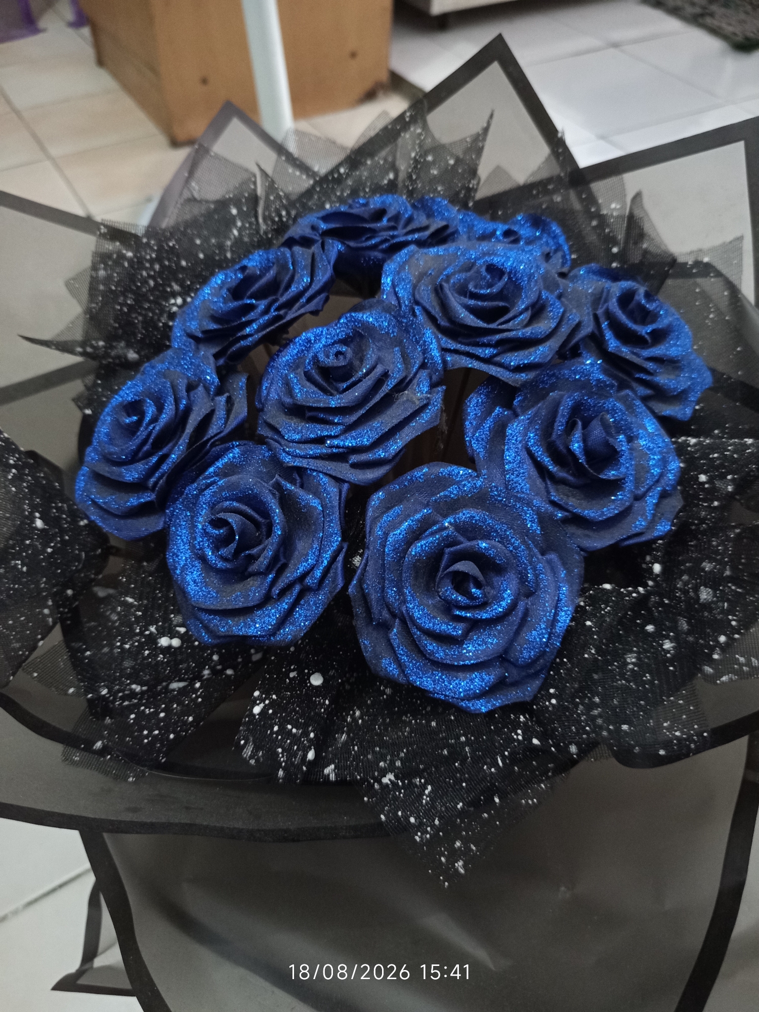 A close-up, indoor, eye-level shot shows a bouquet of approximately twelve artificial, glitter-covered, dark blue roses. The roses are arranged in a circular pattern and are wrapped in black mesh fabric that is speckled with white paint splatters. The bouquet is presented on a dark surface, partially obscuring the base. The background is out of focus, showing a tiled floor and wooden furniture. A timestamp "18/08/2026 15:41" is visible in the lower right corner of the image. The scene is consistent with an indoor setting, possibly a room or a studio, and does not provide any specific visual cues to determine its exact location within Canggu, Indonesia.