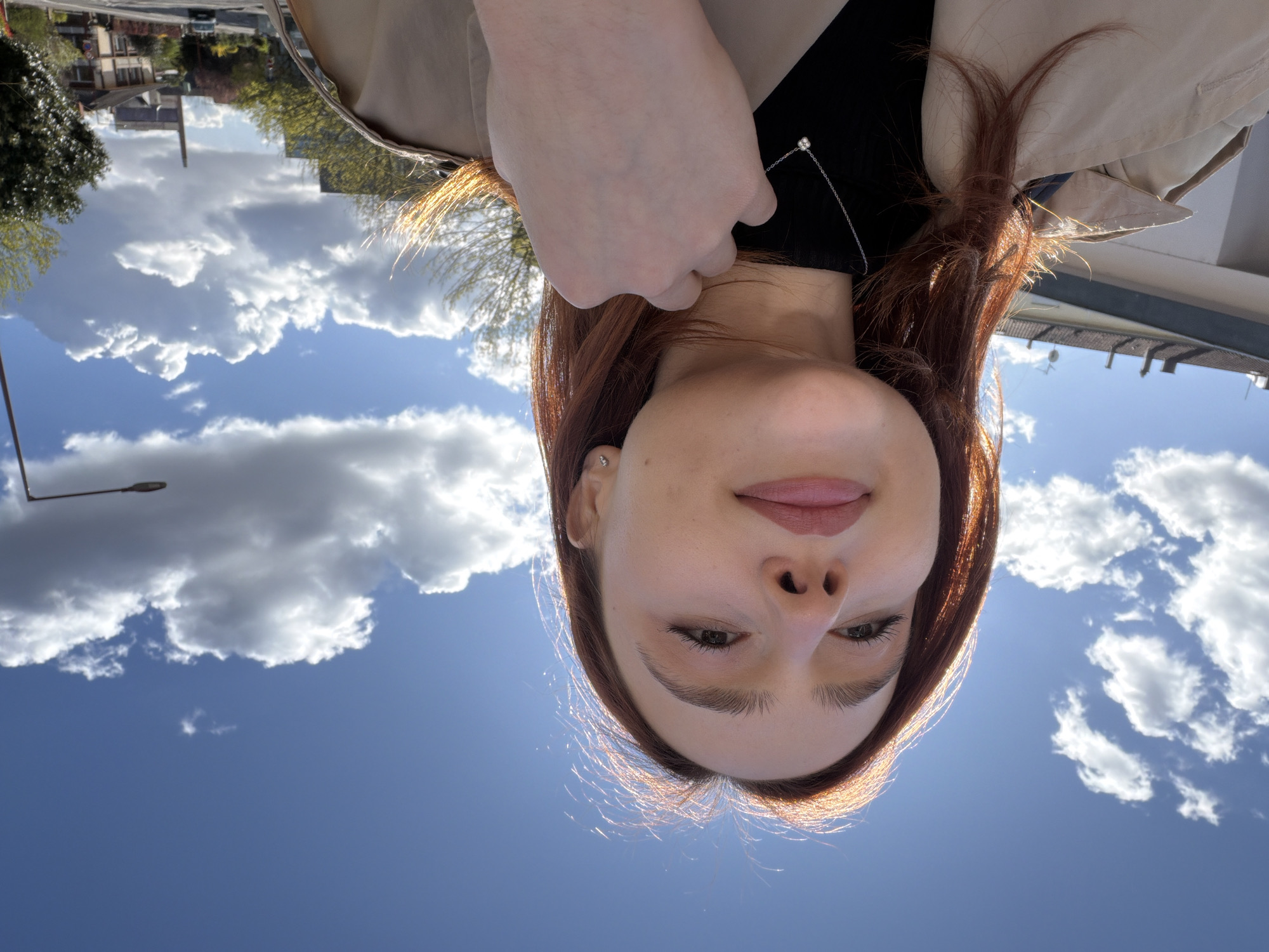 A young female, with reddish-brown hair backlit by direct sunlight creating a halo effect, is depicted in a medium close-up, gazing slightly upwards and to her left. She has light skin, defined eyebrows, and pinkish lip color. She wears a light-colored (beige) trench coat over a black garment, a delicate silver chain necklace with a small clear stone pendant, and small silver stud earrings. Her right hand is positioned at her neck, with fingers slightly curled, touching the collar of her coat.
The scene is outdoors on a bright, sunny day with a deep blue sky featuring scattered white cumulus clouds. In the background, an urban environment is visible. To the upper left, a building with a light-colored facade and a flat or gently sloped roof is present. Further back, and more prominently to the right, a distinctive industrial building with a tall, slender chimney dominates the skyline, identifiable as the `Ancienne Malterie de l'Étoile` (Old Malthouse of Étoile) in the Neudorf district of Strasbourg, France. Residential buildings with pitched roofs, some featuring darker structural elements, line a gently inclining street where a single car is visible. Trees with early spring foliage (some green, some with budding red leaves) are interspersed among the buildings. A dark-colored streetlamp extends from the far right edge of the frame.
