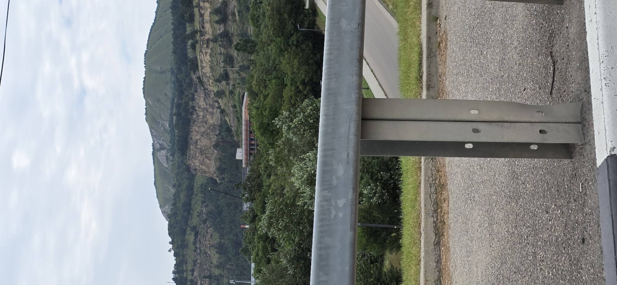 The image captures a view of a hilly landscape with a prominent rock formation and a building under construction, possibly located near Kislovodsk, Russia. In the foreground, a metallic guardrail with support posts is visible, suggesting a roadside vantage point. Beyond the guardrail, a paved road curves upwards into the distance, flanked by grassy slopes. Dense green foliage covers the lower slopes of the hills, partially obscuring a multi-story building with reddish-brown brickwork and visible structural elements, indicative of ongoing construction. A tall, dark crane stands near the building, a common sight at construction sites. The upper parts of the hills are covered in lighter green vegetation, with exposed grey rock formations on the steepest inclines. The sky is overcast with a light, diffused cloud cover, suggesting a temperate or cloudy day. The overall scene is one of a developing urban area encroaching on a natural, hilly environment.