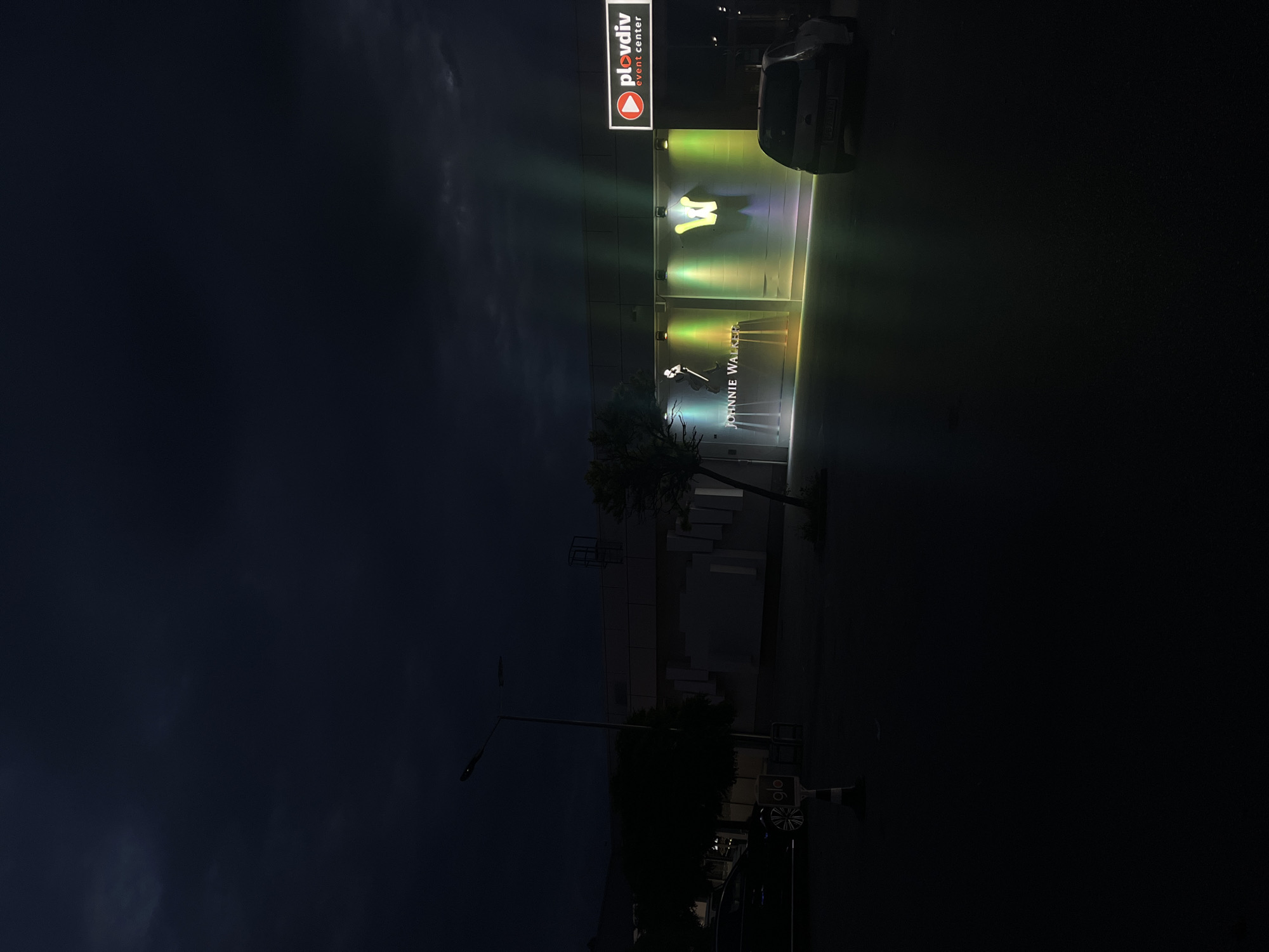 The image captures the exterior of the Plovdiv Event Center at night. The building's facade is illuminated with bright lights highlighting the logos of "Plovdiv event center," "Johnnie Walker," and another logo. A car is parked near the building. The sky is dark, suggesting it is nighttime. A tree stands in front of the building. A street lamp is visible on the left side of the image, along with other parked vehicles.