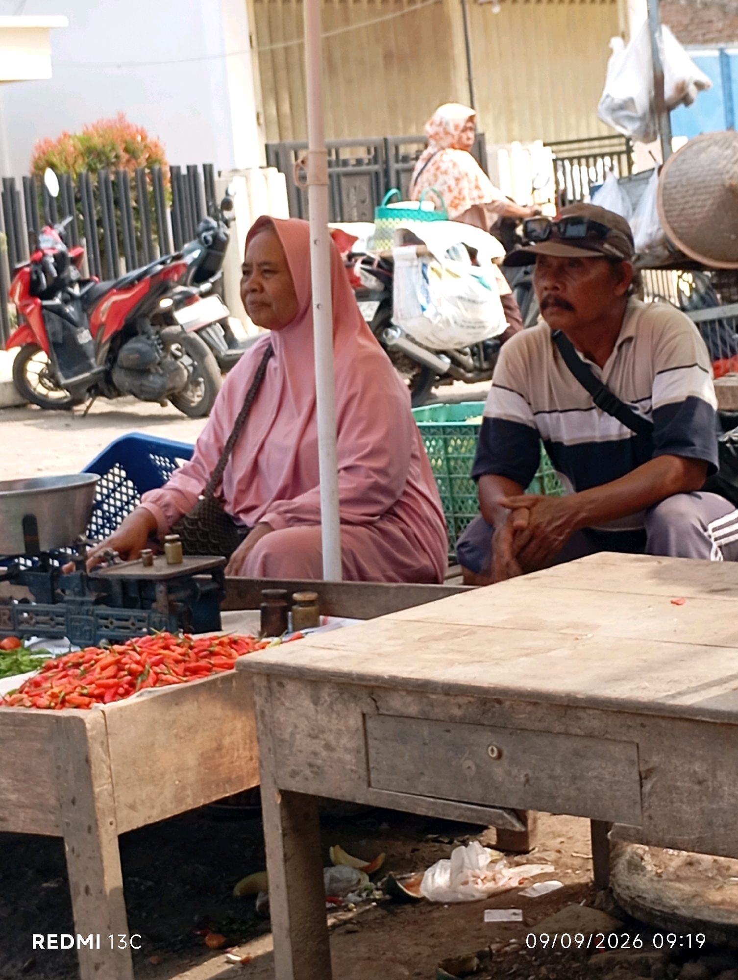 The image depicts an outdoor market scene in Wuluhan, Indonesia, with a focus on a stall selling chilies. In the foreground, a wooden table displays a large pile of red chilies. To the left, a woman in a pink hijab is partially visible, reaching towards a weighing scale. Behind her, a red motorcycle is parked. To the right, a man wearing a cap and a striped shirt is seated, with his hands clasped. Further in the background, another woman in a patterned hijab is visible near a motorcycle. The environment appears to be a street or open-air market, with some debris on the ground. The image is dated 09/09/2026, 09:19.