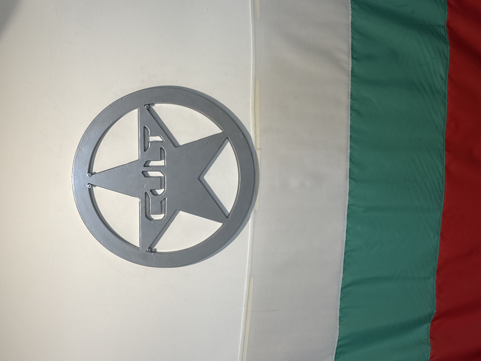 The image shows a flag hanging on a wall. The flag has three vertical stripes of white, green, and red. On the white stripe, there is a gray circle with a five-pointed star inside. The letters "CUП" (CUP) are embedded within the star, seemingly as part of the cutout design.
Without additional context, it is impossible to ascertain the precise location in Varna, Bulgaria.