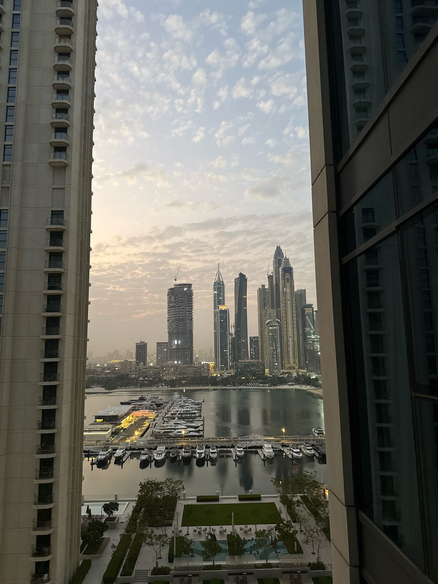 The image captures a waterfront view of Dubai Marina in Dubai, United Arab Emirates, at dusk. The foreground is dominated by the side of a modern residential tower on the left, with a contemporary building's facade and reflective windows on the right. In the middle ground, a marina is filled with numerous yachts moored along docks, with a building complex that appears to be a restaurant or club situated on the water. Beyond the marina, the iconic skyline of Dubai Marina, characterized by its dense concentration of tall skyscrapers, is visible. The sky is partly cloudy, with hues of orange and yellow indicating the setting sun. The overall impression is one of urban development and a vibrant cityscape.
