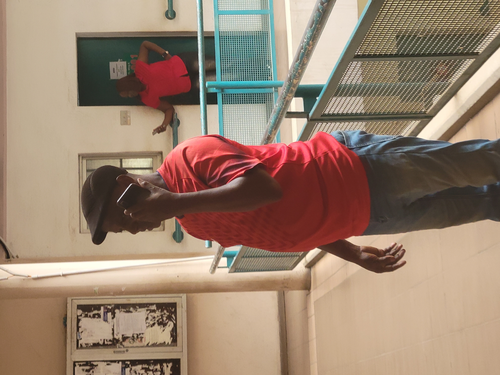 The image, set in an indoor or semi-indoor public building in Abuja, Nigeria, captures two individuals. In the foreground, a man stands, engrossed in a phone conversation. He wears a red short-sleeved shirt, blue jeans, and a dark cap, with a smartphone held to his right ear. His left hand is open, palm facing upwards, at his side.
Further in the background, positioned within an opening in a light-colored wall, another person is visible, also dressed in a red shirt and dark trousers. This individual appears to be leaning forward, possibly observing or interacting with something beyond the frame.
Architectural elements include light-colored walls, a dark green bulletin board behind the person in the background with indistinct white papers, and prominent metal grating structures suggesting a staircase or platform that extends through the middle and upper right of the frame. A gridded window is visible below the foreground man's head. On the bottom left, another bulletin board covered with numerous, unreadable papers is attached to the wall. The scene is illuminated by consistent, bright indoor lighting, indicative of daytime. No specific emotions are evident from the subjects.