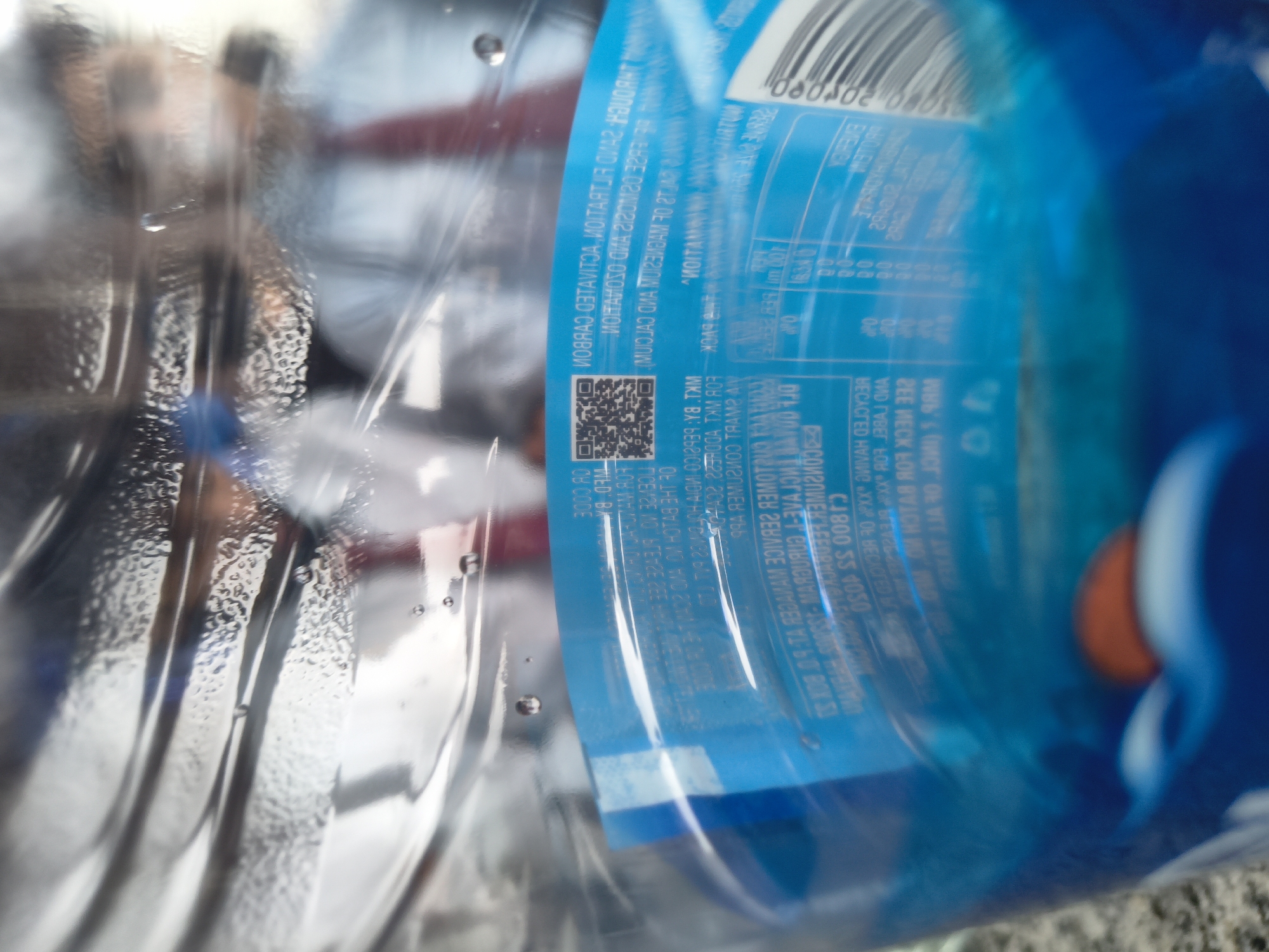 This is a close-up, eye-level, indoor shot looking through a clear plastic water bottle. The bottle is partially filled with water and contains condensation on the interior surface, creating a blurry, distorted view of the background. The background appears to be a dimly lit, indoor environment with reflections of people and possibly office furniture. A blue label with white text, including a QR code, is visible on the lower half of the bottle. The label is partially obscured by condensation and the curvature of the bottle. The image does not contain any information or visual cues that would allow for its location to be identified as Dharamshala, India.