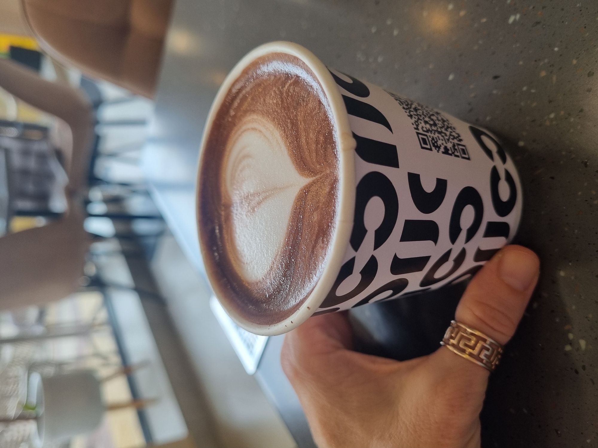 A hand holds a paper cup of latte, which has a visible heart-shaped foam design. The cup is white with black letters and a QR code. The hand has a ring with a geometric pattern on the thumb. The background includes a blurred view of what appears to be a cafe or restaurant interior, including chairs, a table, and a window. The location is specified as Sochi, Russia.