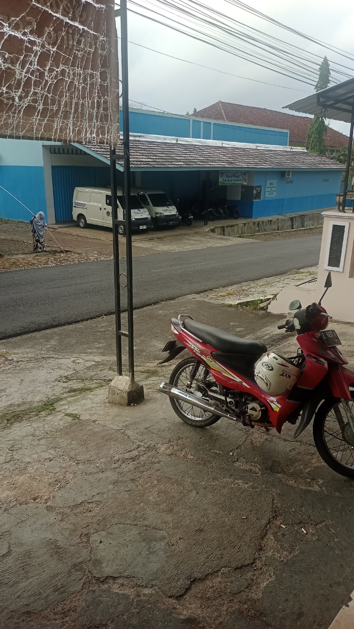 In Karyamandala, Indonesia, a low-angle outdoor scene captures a residential street with a building in the background and a motorcycle in the foreground. The building features a blue facade with white trim and a tiled roof. A white van is parked in front of the building, and several motorcycles are visible on the property. A person dressed in traditional Islamic clothing is sweeping the street. The foreground is dominated by a red motorcycle, partially covered by a white helmet. The ground is paved with concrete. The sky is overcast, suggesting cloudy weather.
