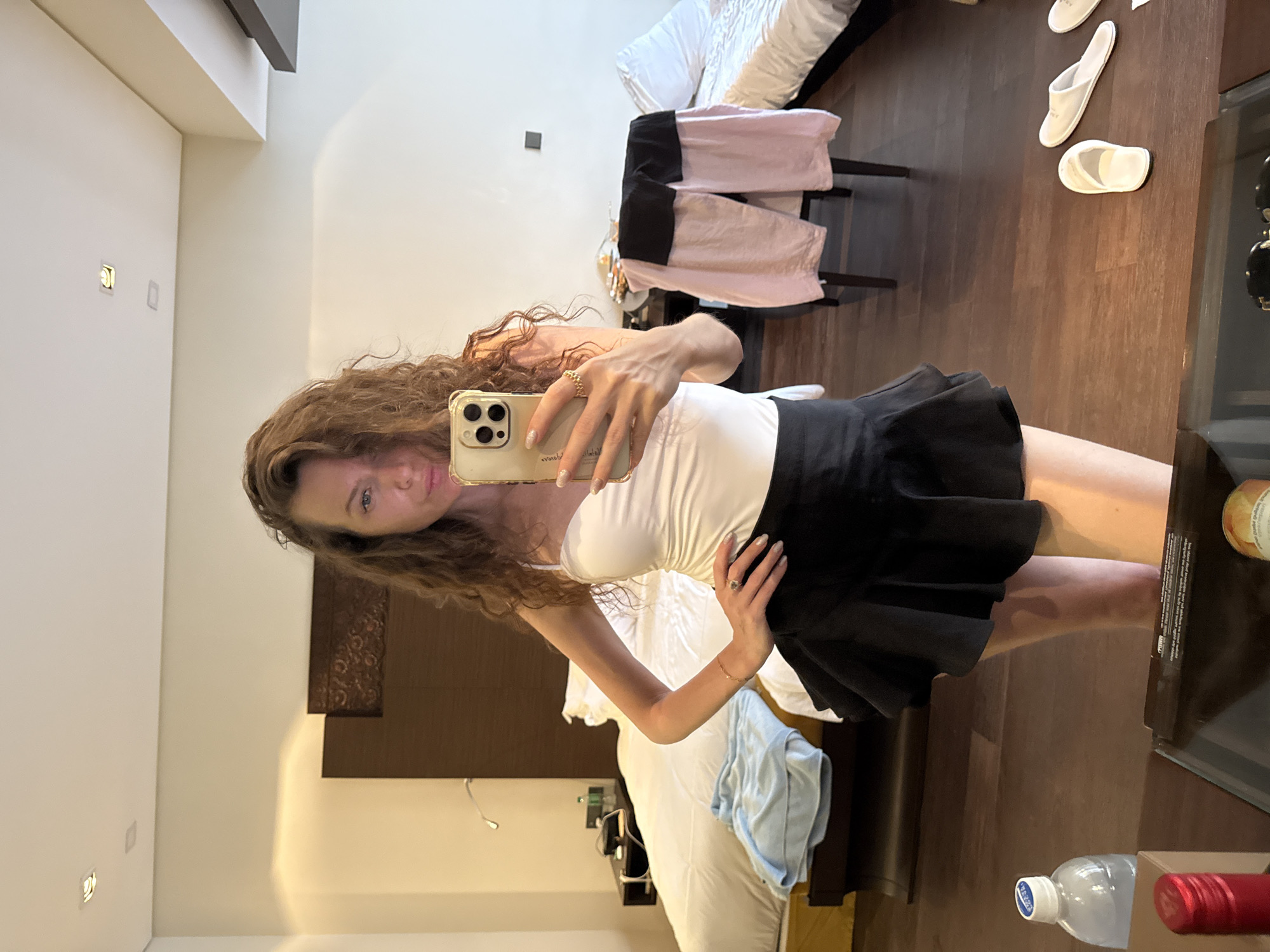 A woman with long, curly brown hair is taking a mirror selfie in a hotel room. She is wearing a white tank top and a black ruffled mini skirt. Her left hand is on her hip, and she is holding a gold-colored smartphone in her right hand. The room appears to be in Dubai, United Arab Emirates, judging by the decor which includes dark wood accents and ornate carvings on the headboard of the bed. The bed is made with white linens and a blue garment is draped over its foot. A chair with pink and black clothing draped over it is to the right of the bed. On the floor in the foreground, there is a bottle of water, and a pair of white slippers.