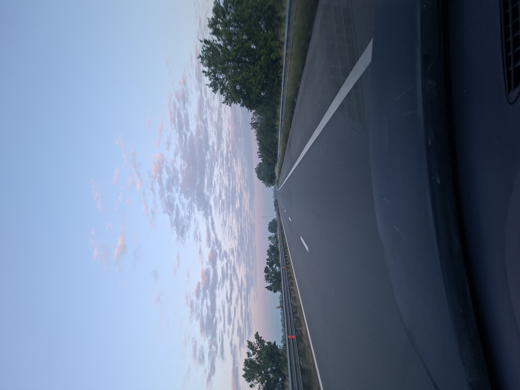 The image shows a multi-lane highway under a sky with light clouds at sunset. The view is from inside a vehicle, looking out across the road. A median with trees runs alongside the highway. There is no visible indication that the scene is in Shishmantsi, Bulgaria. There are no people visible.