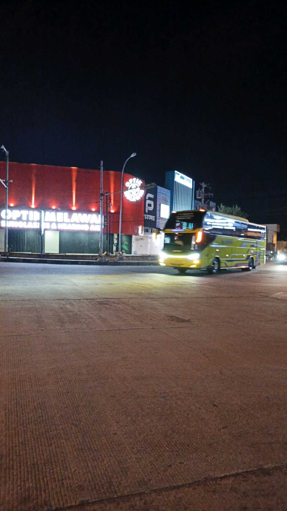 In the city of Surakarta, Indonesia, at night, a yellow bus is captured in motion on a street. The bus is illuminated by its headlights and interior lights. To the left of the bus is a building with a red facade and large white lettering that reads "OPTIN MAILAWAI". Above the lettering, a circular logo with a stylized "P" is visible. A smaller building with a blue facade and "STORE" written in white lettering is to the right of the red building. The street appears to be made of concrete with visible lines. The sky is dark, indicating it is nighttime.