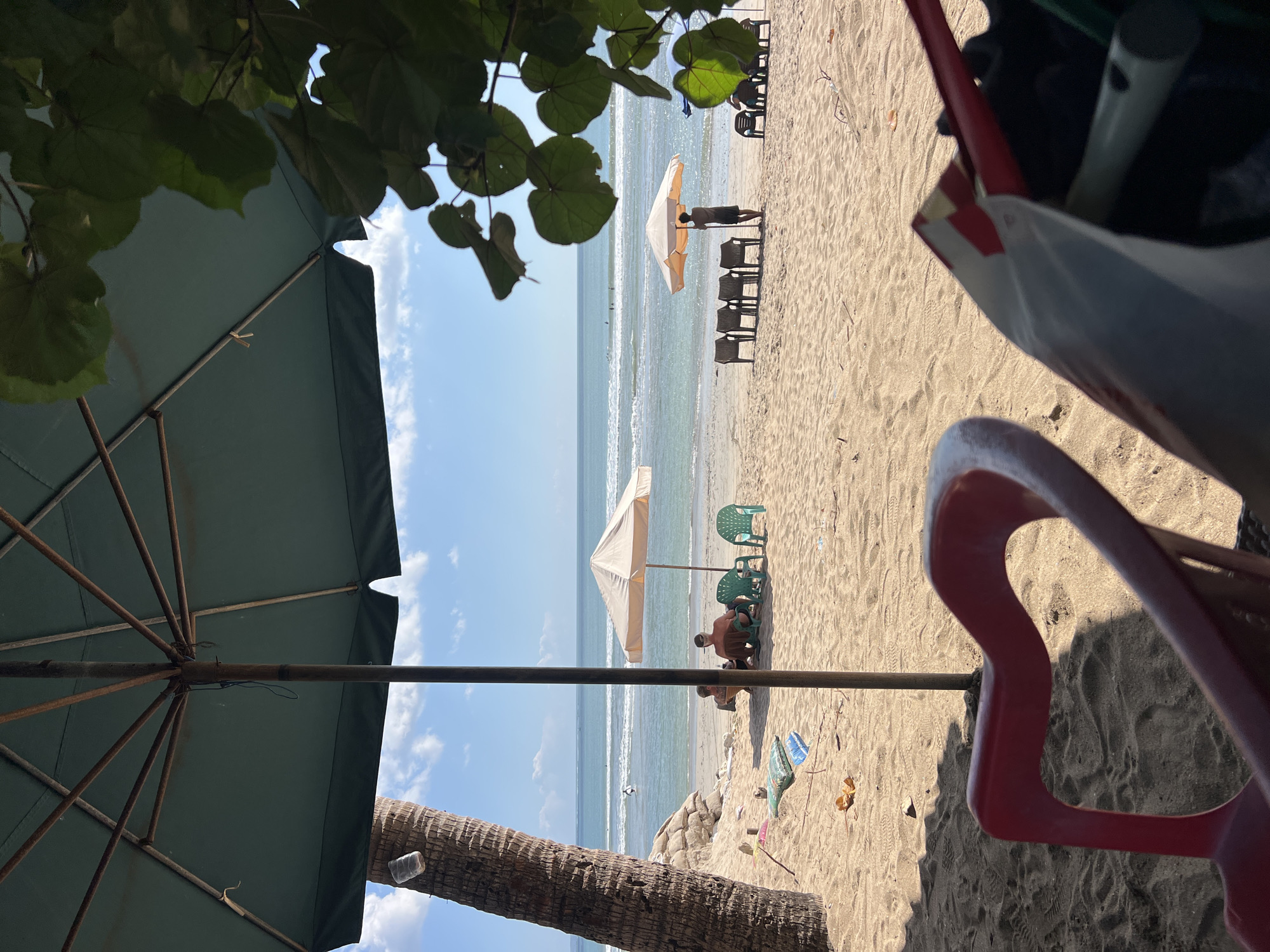 A beach scene unfolds in Kuta, Indonesia, under a clear blue sky dotted with white clouds. In the foreground, the edge of a red chair and a bag are visible, suggesting a seated perspective. A large green umbrella, supported by a wooden pole, extends over the scene, with its spokes visible. To the left, a palm tree stands tall, its trunk rough and textured.
Further into the scene, a sandy expanse stretches towards the ocean. Several beach umbrellas are set up. One light-colored umbrella shades a couple sitting on chairs. Another umbrella, further to the right, has a person standing beneath it, seemingly arranging chairs. In the middle distance, a line of empty chairs awaits beachgoers. The ocean appears calm, with gentle waves rolling towards the shore. A few distant figures can be seen in the water, some appearing to be surfing. The sand itself is textured with footprints and scattered debris, including a plastic bottle tied to the palm tree and some colorful objects near the water's edge. The overall atmosphere is relaxed and typical of a tropical beach setting.