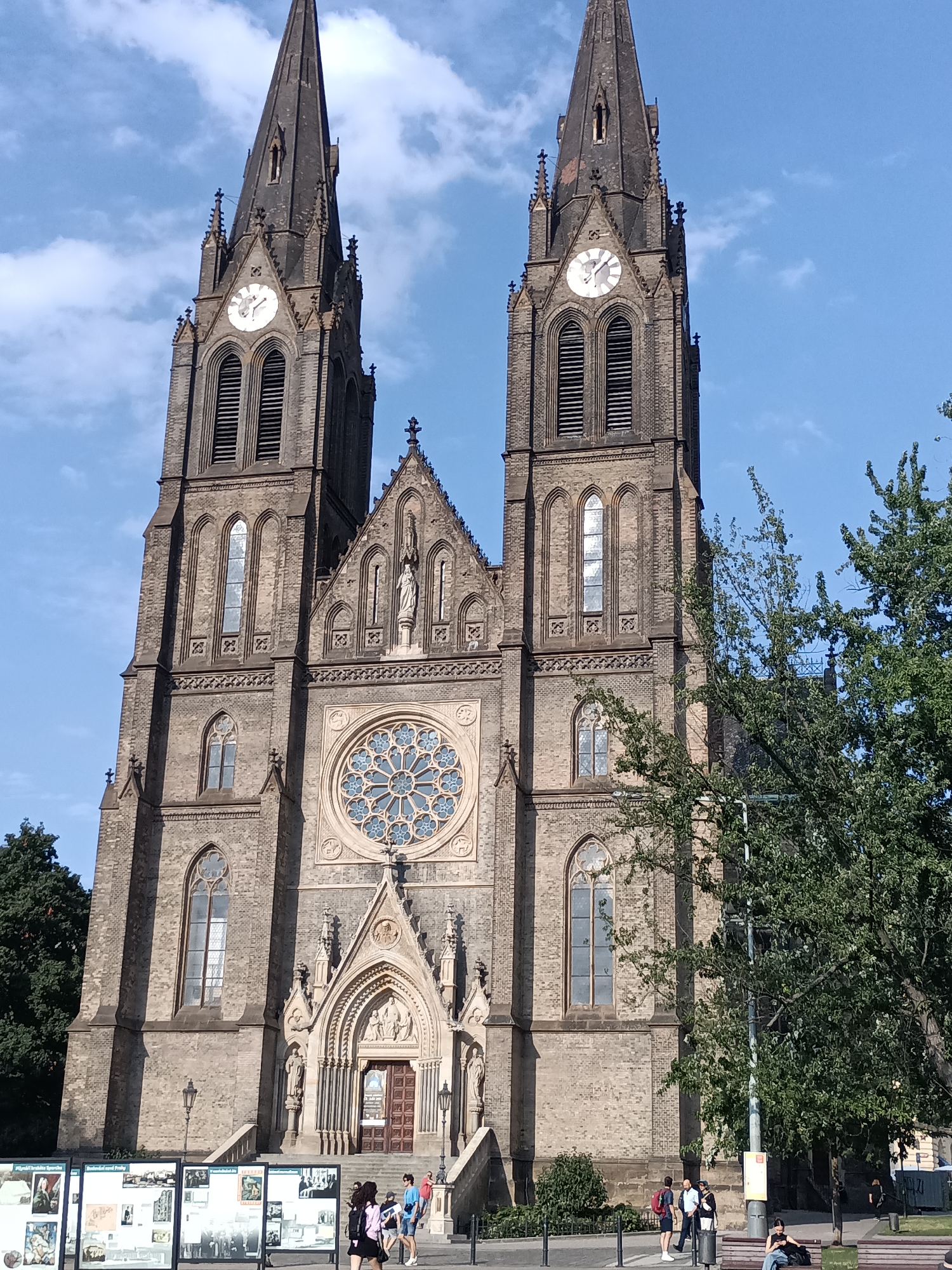The image shows a frontal view of the Basilica of Saint Peter and Saint Paul in Vyšehrad, Prague, Czechia. The basilica is a prominent Gothic Revival structure with two tall spires, each featuring a clock face. A large rose window is centered on the facade above the main entrance, which is adorned with sculptures and Gothic architectural details. The sky is blue with scattered white clouds. In the foreground, there are several display boards with images and text, and a few people are walking and standing around the entrance. Green trees partially obscure the right side of the building. The scene appears to be a sunny day.