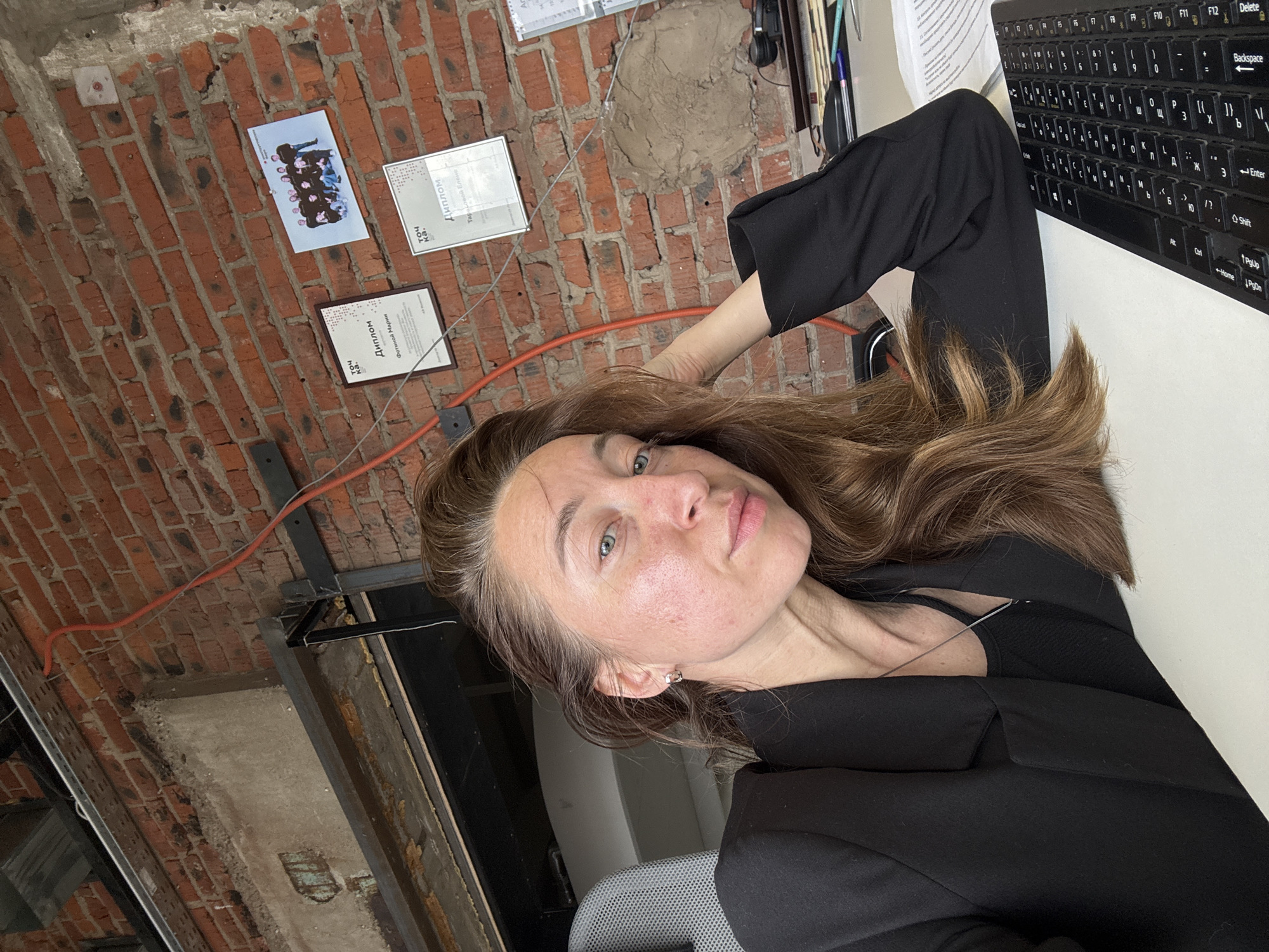 The image shows a woman captured from a slightly upward, tilted perspective, set in an indoor environment in Chaikovsky, Russia. She has long, wavy brown hair, light-colored eyes, and a neutral, direct gaze towards the camera. She is wearing a black blazer over a dark top, with her right arm bent and her hand near the side of her head.
The background is dominated by a raw, exposed red brick wall, which appears diagonally in the frame due to the image's orientation. An orange electrical cable runs across the brick, and some patches of concrete or plaster are visible. Attached to the brick wall are two framed diplomas or certificates; one clearly displays "Диплом" (Diploma) and "Фотиной Марии" (Fotinoy Marii), along with "точ ка." (toch ka.), while the other also shows "Диплом" and partially "Тар". A small, printed photograph of a group of people is also pinned to the wall.
To the right, a section of a white desk surface is visible, holding a black computer keyboard. The keyboard features both Latin and Cyrillic characters, with keys such as "Delete", "Backspace", "Enter", "Shift", "Ctrl", "PgUp", "Home", and "PgDn" clearly discernible. Some papers with text are partially obscured behind the keyboard. The lighting suggests an indoor setting, likely an office or creative workspace, characterized by its industrial-chic aesthetic with exposed brick.