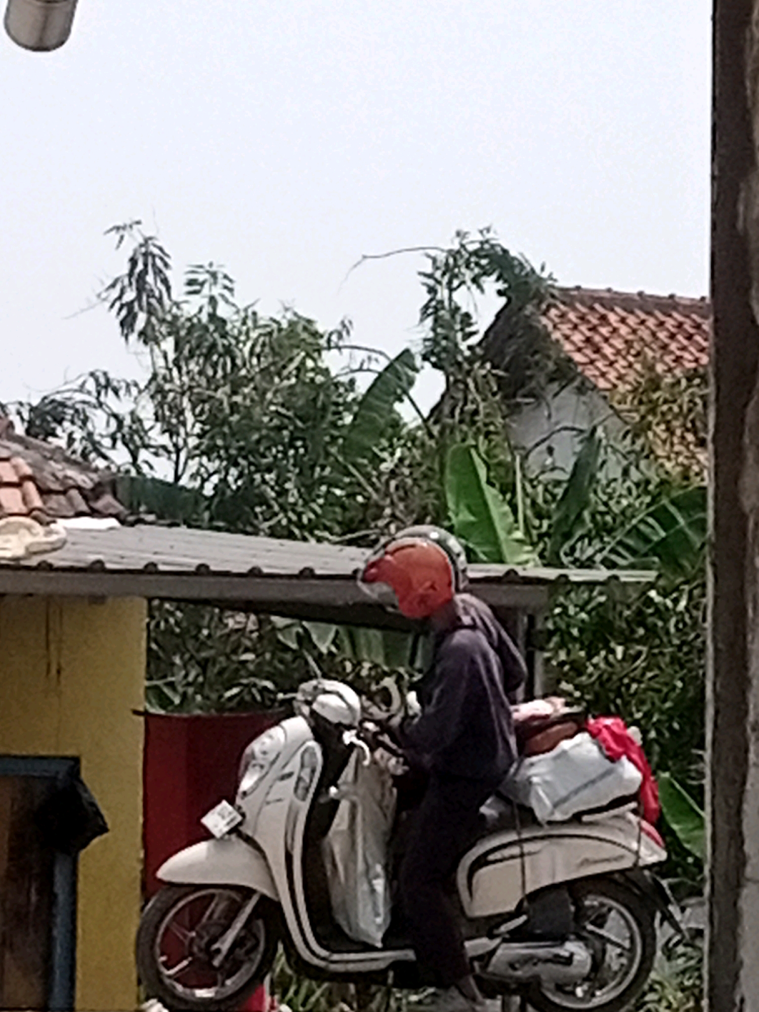 A person wearing a dark purple hoodie and an orange helmet is sitting on a white scooter in what appears to be an outdoor setting. The scooter is loaded with bags, including a white bag and a red piece of fabric. The background shows green foliage, rooftops, and a cloudy sky. The scene is likely located in Boyoteluk, Indonesia, as indicated by the provided context. The overall impression is of a person preparing to travel or having just arrived.
