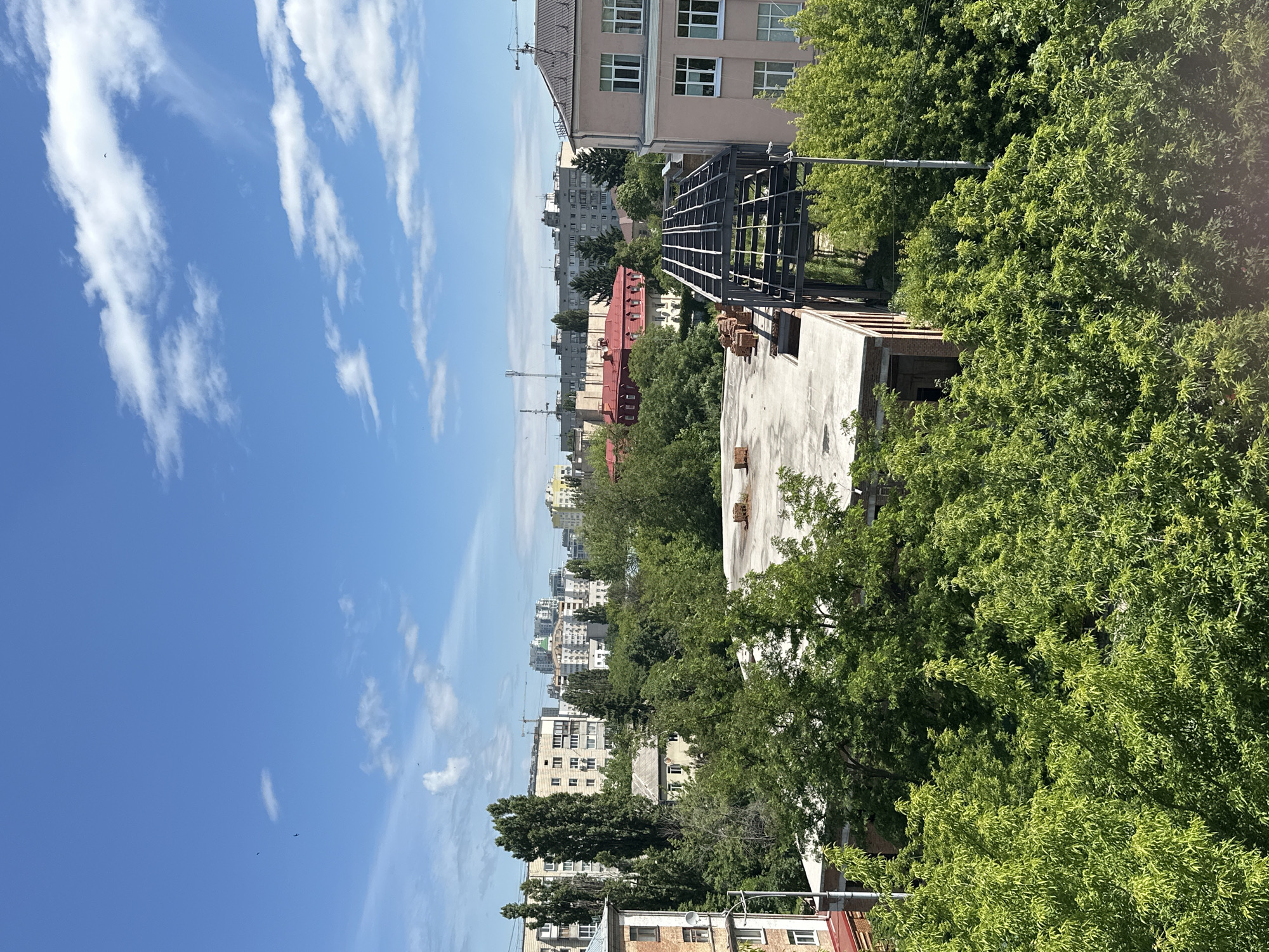 The image captures an urban landscape in Kyiv, Ukraine, on a clear, sunny day with scattered clouds. The foreground is dominated by lush green trees, some of which partially obscure buildings. In the mid-ground, several buildings are visible, including a flat-roofed structure with exposed bricks and a partially constructed metal framework on its roof. Further back, a mix of apartment buildings and other structures form the city skyline. Some buildings are modern, while others appear older. The sky is a vibrant blue, with thin white clouds drifting across. Several small birds are visible in the sky. The overall impression is a typical urban scene on a bright day.