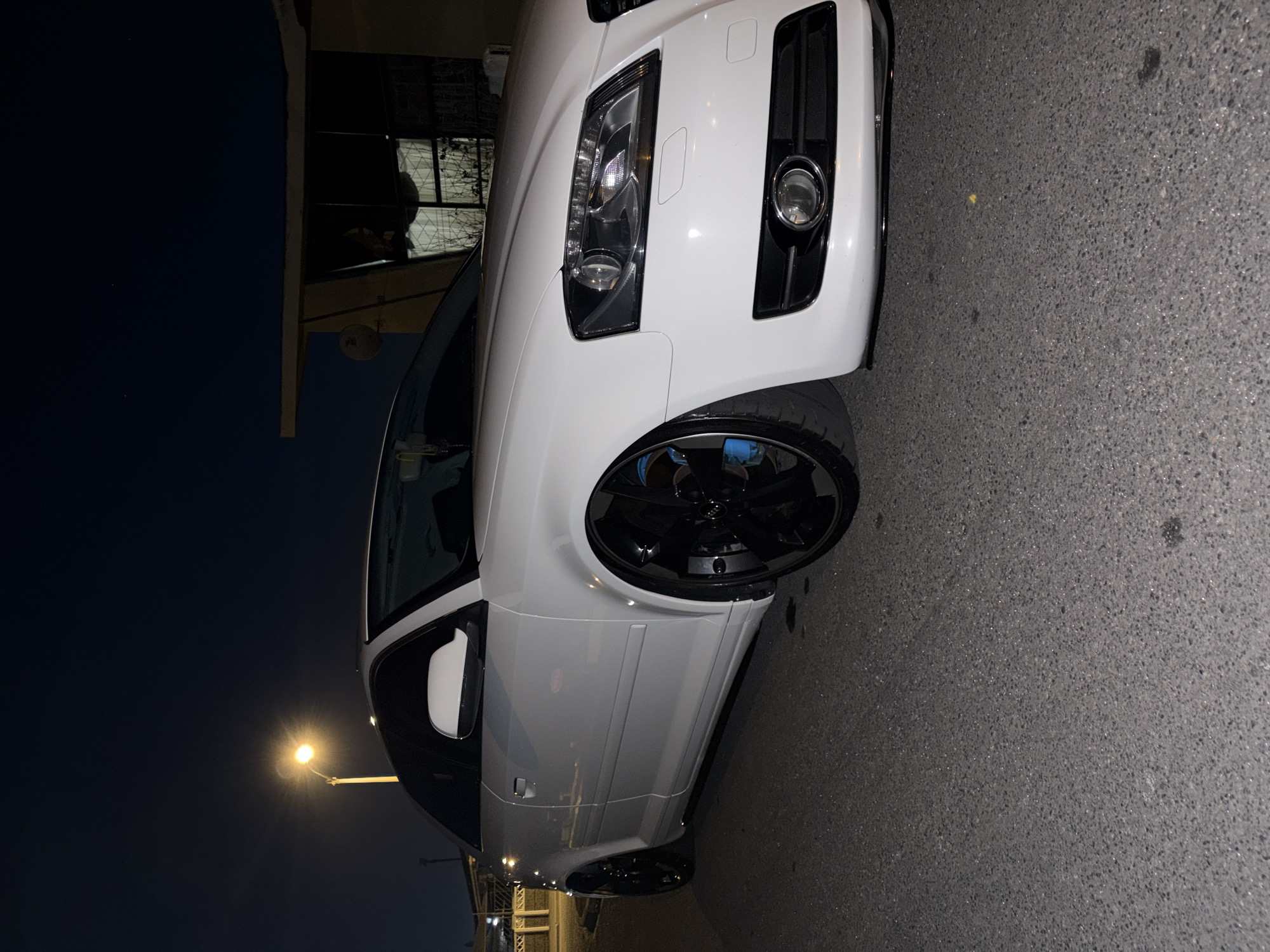 A white Audi A3 coupe is parked on an asphalt road at night. The car has black rims and bright blue brake calipers. In the background, a streetlamp illuminates the scene, and a portion of a building with a satellite dish is visible. The image appears to be taken in Goodwood, South Africa, judging by the surrounding context. There are no people visible in the image.
