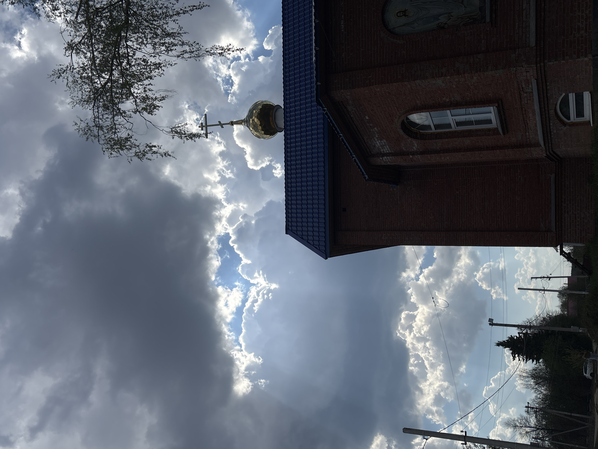 The image shows an upward-angled view of a red brick building with a blue tiled roof and a golden onion dome topped with a cross, likely an Orthodox church. A tree branch appears at the top left. The sky is visible behind the building, featuring a mix of dark storm clouds and bright white clouds. A window and religious art are visible on the building's facade. In the distance, utility poles and trees are present. The location is Фоки, Russia.