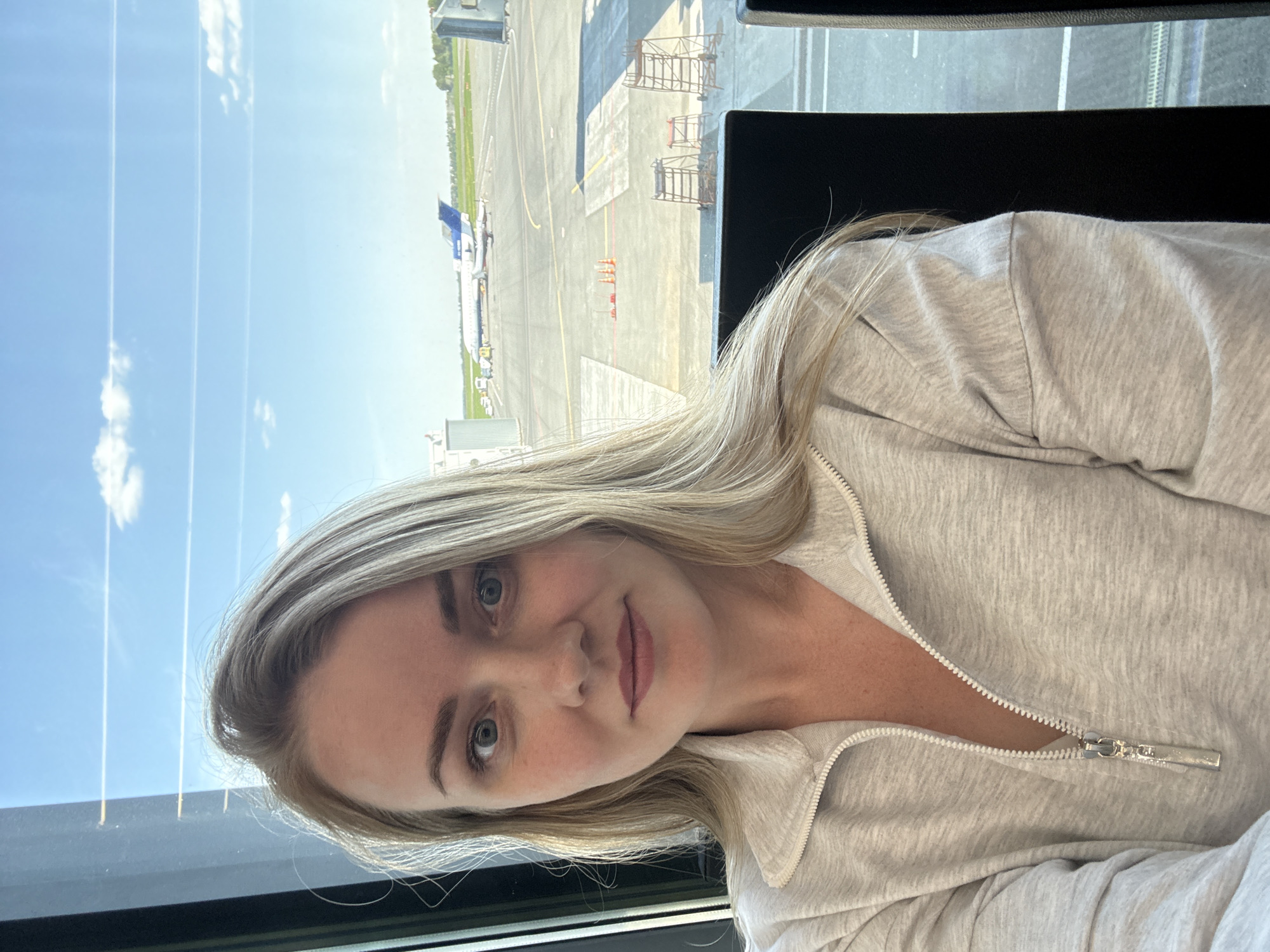 A woman with blonde hair and fair skin is posing for a photo in what appears to be an airport terminal. She is wearing a light gray zip-up sweater. Outside the window behind her, an airplane is visible on the tarmac. The environment includes a clear blue sky and airport infrastructure. The location of the scene in the city of Новое Мартьяново, Russia cannot be verified from the available visual information.