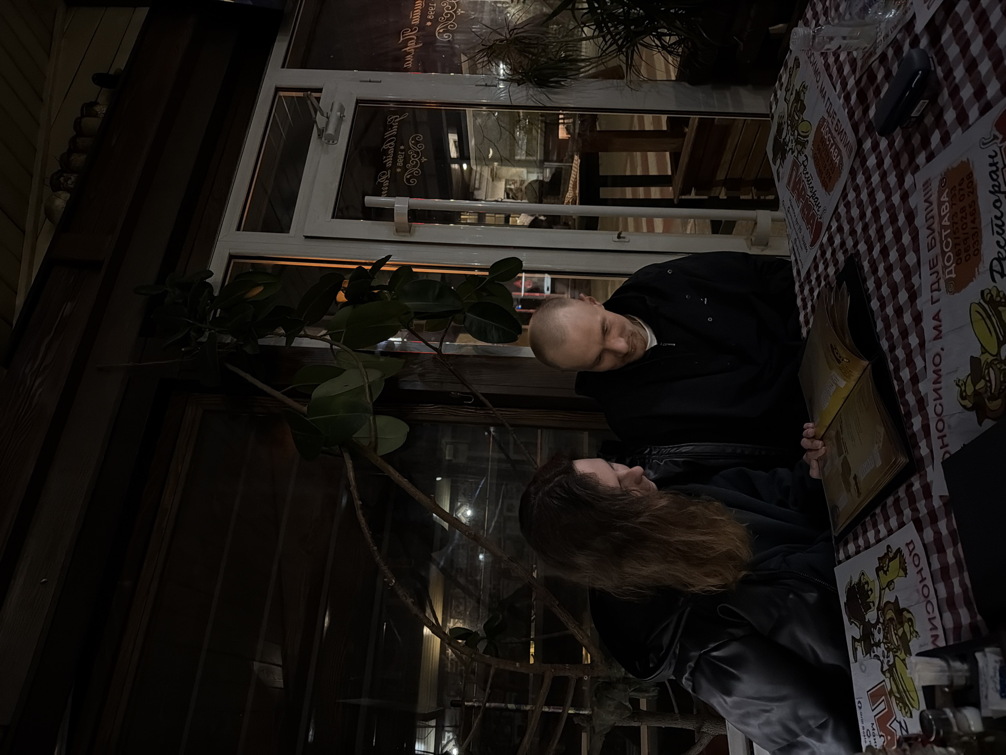 Here is a forensic description of the image:
**People:** Two individuals are present: a man with a bald head and a woman with long, wavy hair. Both are wearing dark clothing.
**Subjects:** The two individuals appear to be dining at an outdoor restaurant. They are looking at a menu together.
**Objects:** A restaurant table covered with a checkered tablecloth displaying menus and what appears to be promotional material in Serbian. The menu has an image of a cartoon chef on it. A glass window behind them shows the restaurant's interior. There is a plant visible to the left of the window.
**Actions:** The man and woman are looking at the menu.
**Interactions:** The individuals appear to be engaged in the activity of selecting what they want to eat.
**Environment:** The setting appears to be an outdoor patio of a restaurant at night. Given the text on the menus in Serbian, this would likely be in Budva, Montenegro.