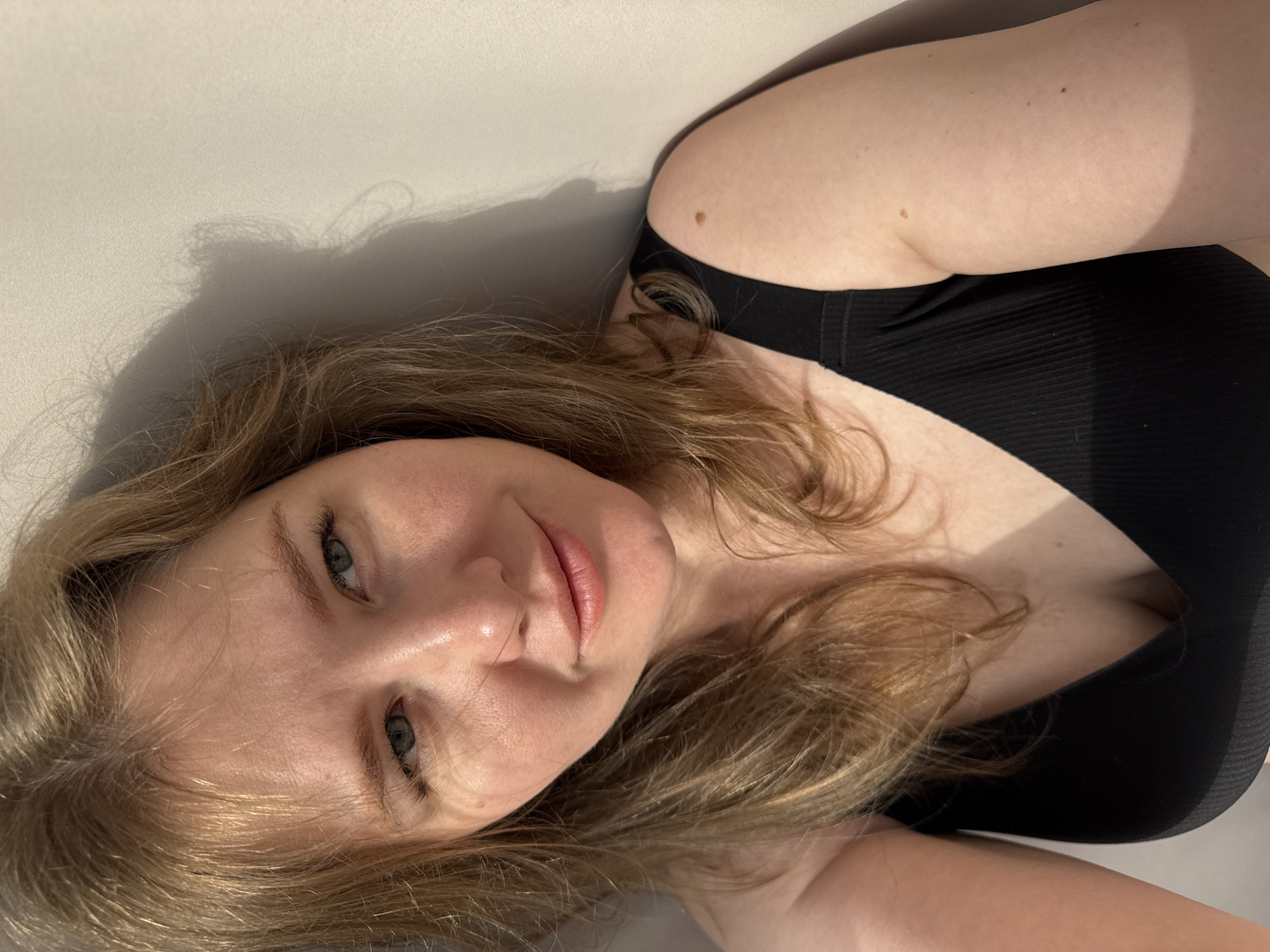 A young woman is depicted lying down, with the camera positioned above her, capturing a close-up, angled shot. Her head is turned slightly, and her light-colored eyes (blue or green) look directly at the viewer. She has long, light brown to blonde hair that spreads around her head and shoulders. Her lips are a soft pink, and her expression is neutral to serene.
She is wearing a black, ribbed top or bra, part of which is visible across her chest and shoulder. Her skin appears fair, with a few small moles visible on her arm and shoulder. Strong, directional light casts prominent shadows behind her head and body onto a plain, light-colored surface, likely a wall or bed. This lighting suggests an indoor setting, possibly illuminated by sunlight through a window during the daytime. There are no visible objects or text within the frame, and the background is minimalist, focusing all attention on the subject. The overall impression is an intimate portrait, capturing a moment of quiet repose.