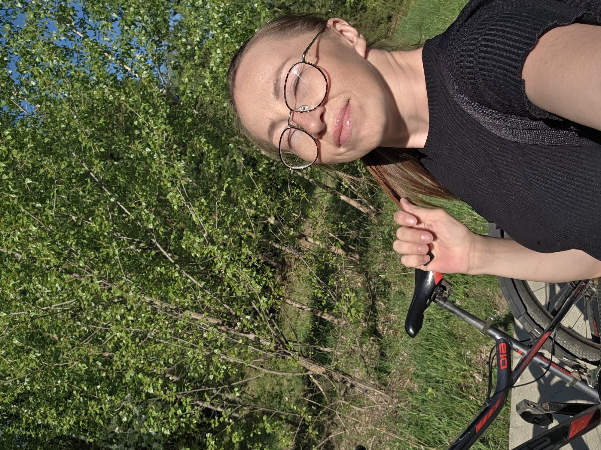 A woman with round glasses is standing outside with a bicycle. She has light skin and light brown hair. She is wearing a black, textured shirt. The bicycle is black and gray with red accents. There is a dense background of green trees and grass. The location is Chaikovsky, Russia.