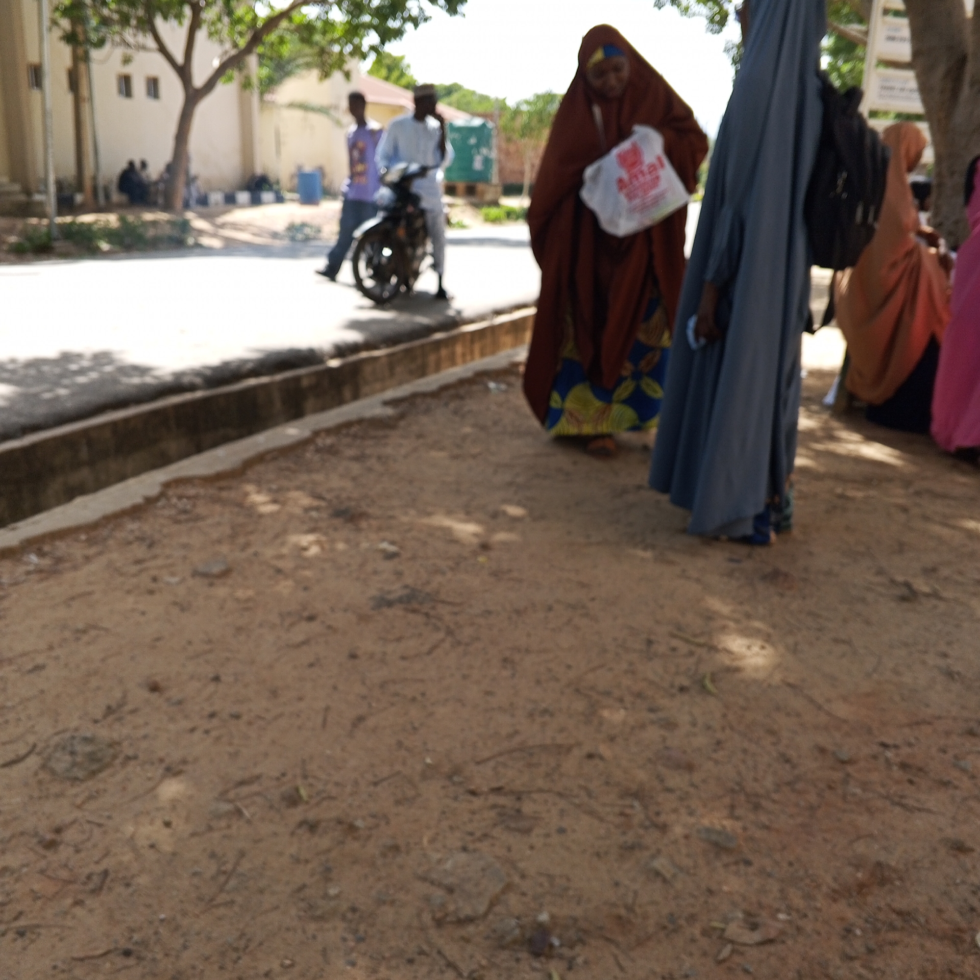 In Gombe, Nigeria, on a dirt road with a concrete drainage ditch to the left, a group of people are gathered. In the foreground, the dusty ground is sparsely littered with small debris. To the left, a concrete curb runs along the edge of a paved road, which is in shadow. Behind the curb, a few people are visible, including one on a motorcycle, another walking, and two others sitting. Further back, a building with two windows is visible, with a tree partially obscuring it. The main focus of the image is a woman in the middle ground wearing a brown abaya and a headscarf, holding a white bag with red writing. To her right, another person is wearing a grey abaya and a black backpack. To the right of the frame, two other individuals are partially visible, one in an orange hijab and another in a pink garment. The scene suggests a casual gathering or movement of people on a street.