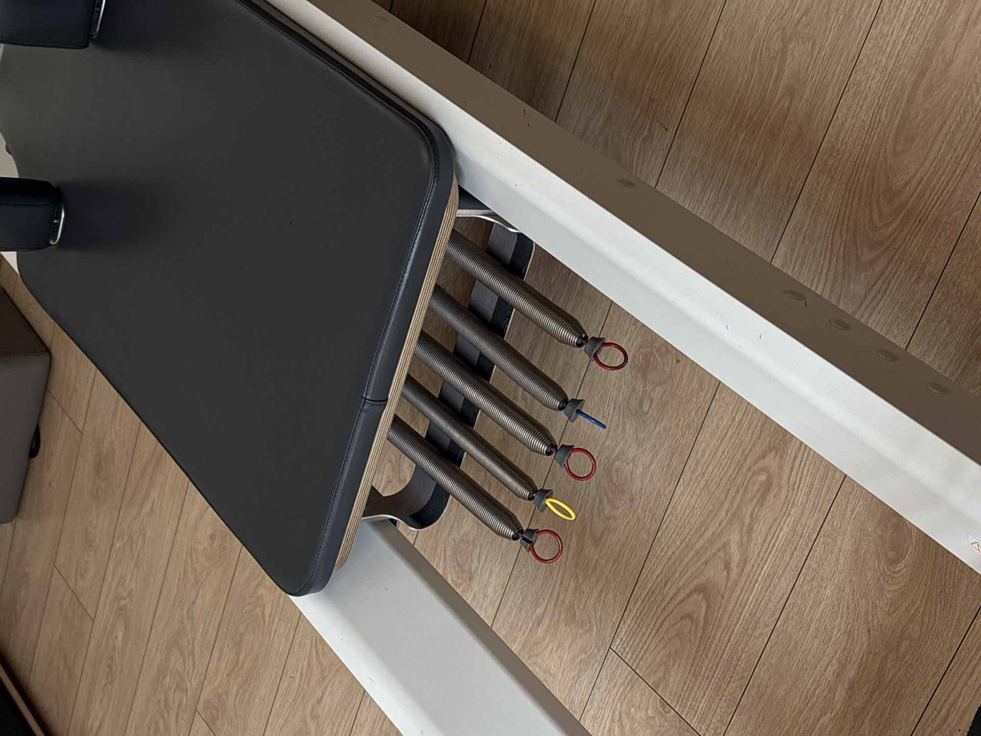 The image provides a close-up, angled view of a modern Pilates reformer situated on a wooden floor. The primary subjects are the components of the fitness equipment and the flooring.
In the foreground and central to the image is the black, padded carriage of the Pilates reformer, seen from an elevated perspective. The carriage has a sleek, rectangular shape with visible stitching along its edges. Beneath the carriage, a light-colored wooden platform is partially visible.
To the right of the carriage, four metal resistance springs are prominently displayed. Each spring is attached to the reformer's frame and features a distinctively colored fabric loop at its end. From bottom to top, these loops are red, yellow, red, and blue, typically signifying different resistance levels. The springs connect to a dark grey or black mechanism that is part of the reformer's structure.
The reformer itself has a sturdy white frame that runs diagonally across the right side of the image. The floor is made of light brown wooden planks with noticeable grain patterns, suggesting an indoor setting like a Pilates studio or home gym.
No people are visible, and no specific activity is taking place; the equipment appears stationary. There is no visible text within the image. The time of day and weather are not discernible, as the scene is indoors. While the context of Mexico City, Mexico is provided, there are no specific visual cues within the image itself to confirm this location.