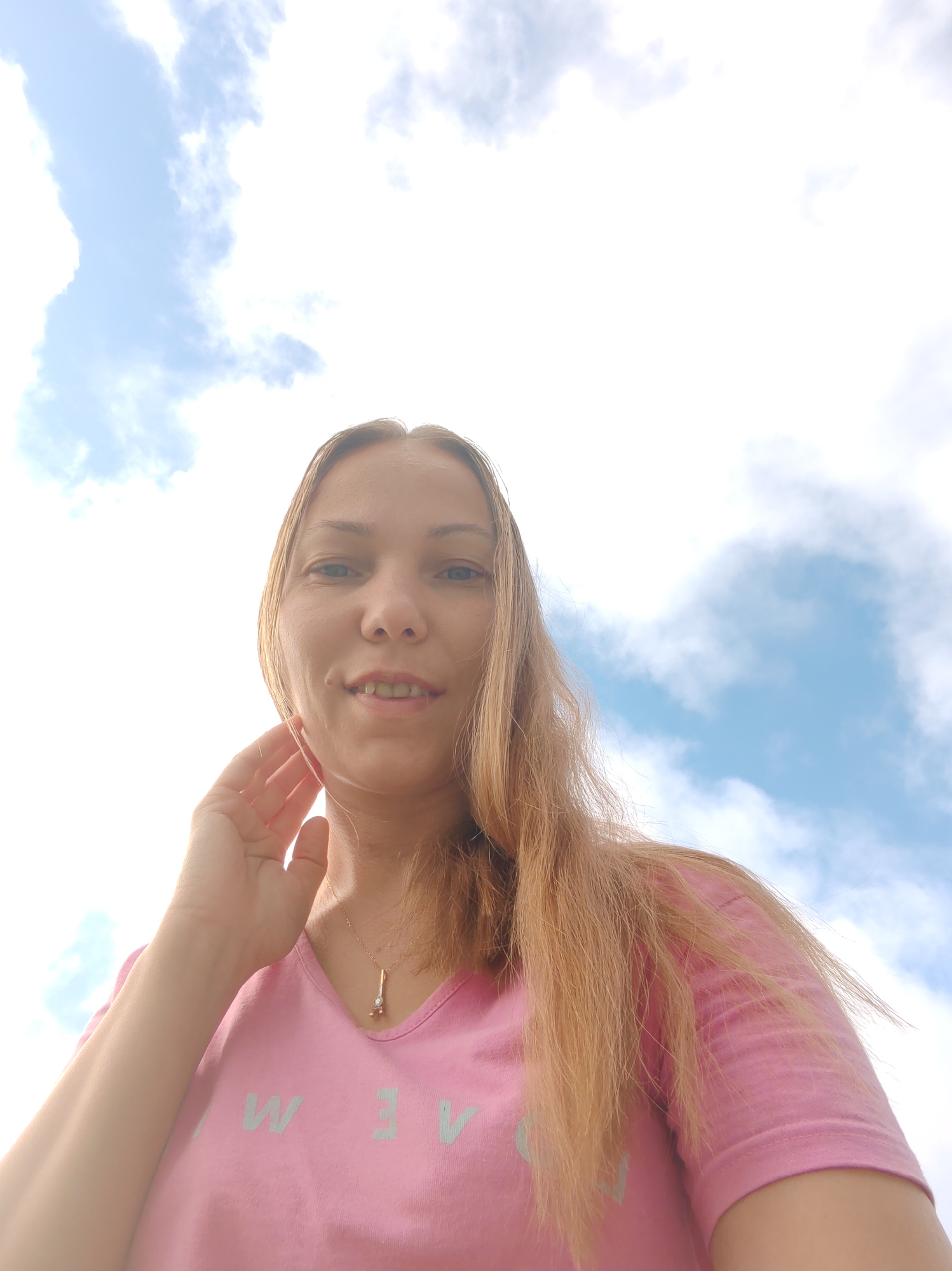 The image shows a young woman with long, light brown hair and fair skin, looking up at the camera. She is wearing a pink t-shirt with a light blue graphic design that appears to be text. Around her neck, she wears a delicate gold necklace with a small pendant. Her left hand is raised to her cheek. The background is a bright blue sky with scattered white clouds, suggesting an outdoor setting. The perspective is a low-angle shot, making the sky a dominant feature. The image appears to be a selfie taken on a sunny day, possibly in a park or open area within the city of Chuhlem, Russia, though no specific landmarks are visible.