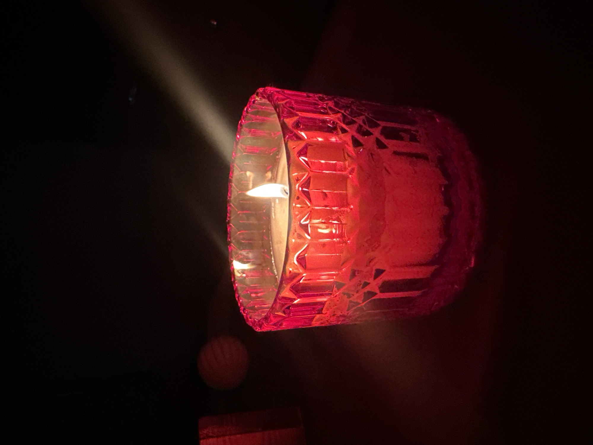 A lit red candle in a textured glass holder sits on a dark surface. The candle flickers with a bright flame. To the left of the candle, partially visible, is a red rectangular object. In the background, a beam of light cuts through the darkness. The overall scene is dimly lit and appears to be indoors, with no discernible elements suggesting a specific location within the city of Kharnavy, Russia.