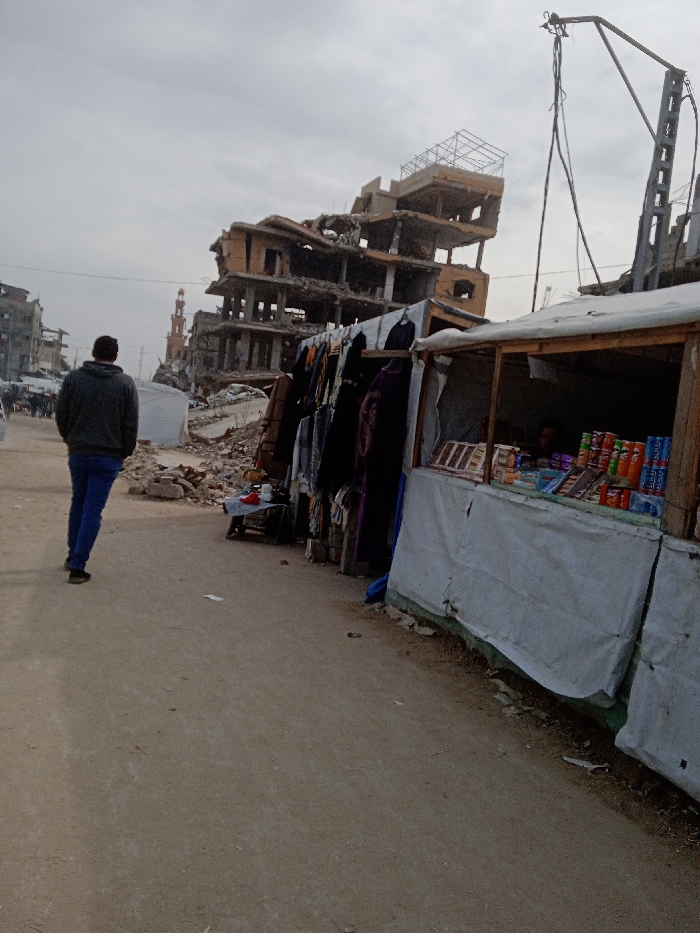 The image displays a street scene in Khan Yunis, Palestinian Territory, under an overcast sky. A light-colored dirt road or pathway constitutes the foreground. On the left side of the path, a male individual, dressed in a dark gray hooded sweatshirt and blue jeans, walks away from the camera. To the right, two makeshift market stalls are present. The stall closer to the camera features a wooden frame and white tarpaulin coverings; its interior displays rows of packaged snacks, including chip cans and juice boxes, with a person partially visible inside. Adjacent to this, another stall presents various dark-colored clothing items, resembling robes and dresses, hanging for sale.
The background is dominated by severely damaged multi-story concrete buildings, exhibiting widespread structural collapse, exposed rebar, and debris. One prominent building on the left appears extensively destroyed. Further in the distance, behind the damaged structures, a light-colored mosque minaret with a pointed top is visible. On the far right of the frame, a damaged utility pole-like structure with hanging wires extends upwards. The scene depicts a street in a post-destruction environment, where improvised commerce is occurring amidst significant urban damage.