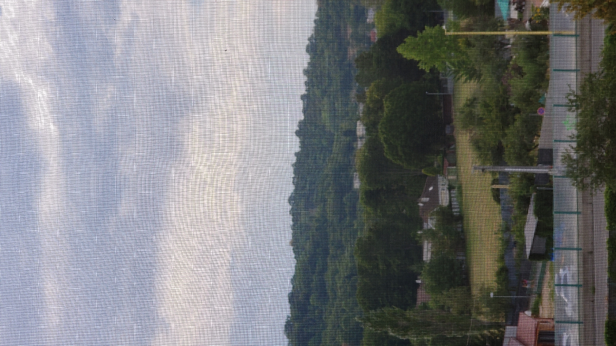 This image, captured through a window screen, displays a suburban landscape under a cloudy sky. In the foreground, a road is visible, bordered by a green fence. Beyond the fence, there are scattered houses, some with red roofs, and various trees. A grassy field with some markings is situated behind the houses. In the middle ground, a densely wooded hill forms the backdrop, with some building rooftops peeking through the trees. The sky above is overcast, with patches of brighter clouds suggesting diffused sunlight. The scene appears to be a residential area in Prešov, Slovakia, characterized by a mix of residential buildings, natural greenery, and infrastructure.
