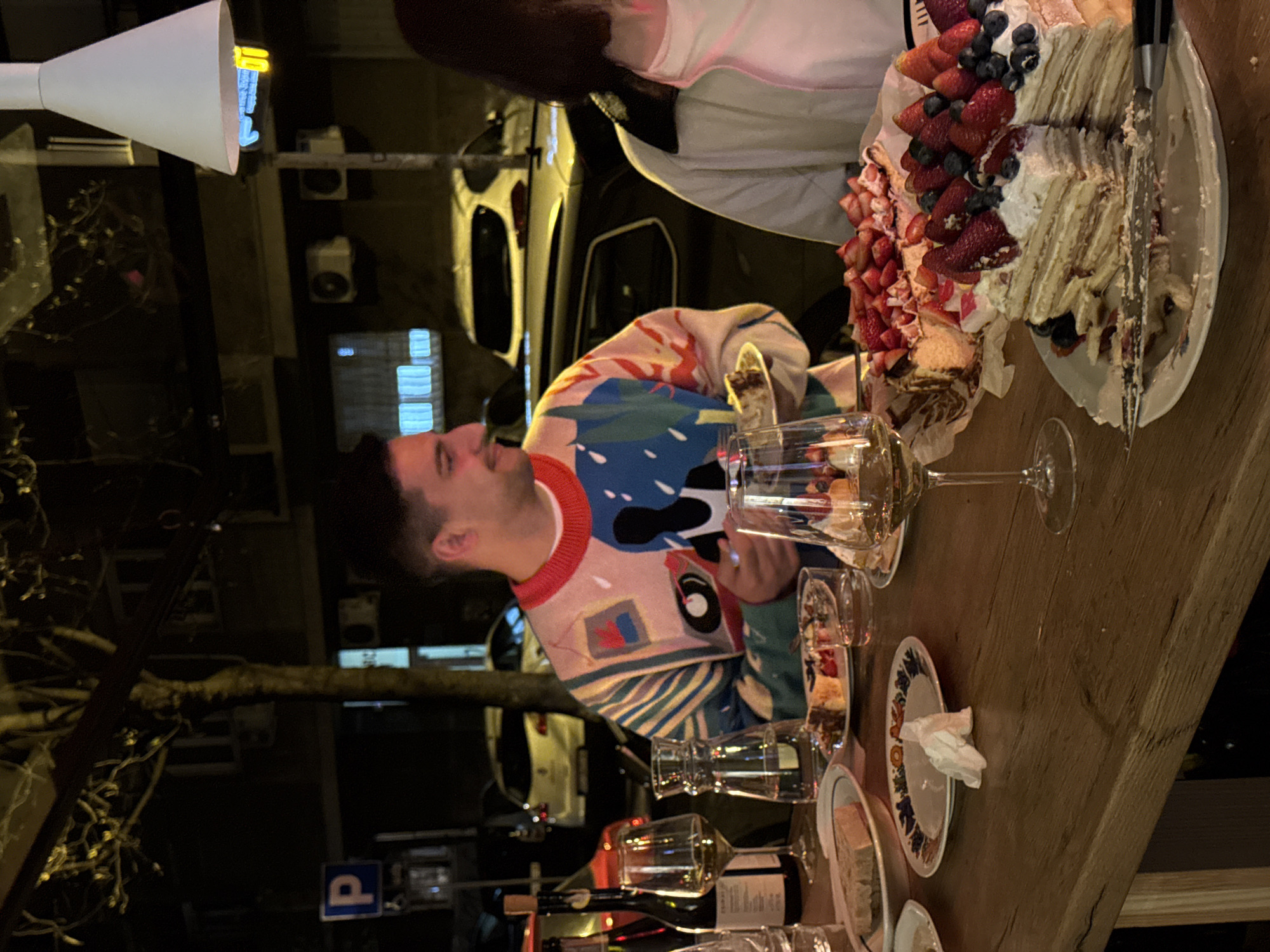 A man in a colorful patterned sweater is seated at a wooden table outside, seemingly enjoying a meal or gathering. Next to him is a large cake decorated with strawberries and blueberries. The table is set with glasses, plates, and wine bottles, indicating a social setting. The location appears to be in Belgrade, Serbia, as suggested by street signs and architectural features. Cars are parked in the background, and there are trees along the street. The scene is captured at night, with artificial lighting illuminating the table and surrounding area.