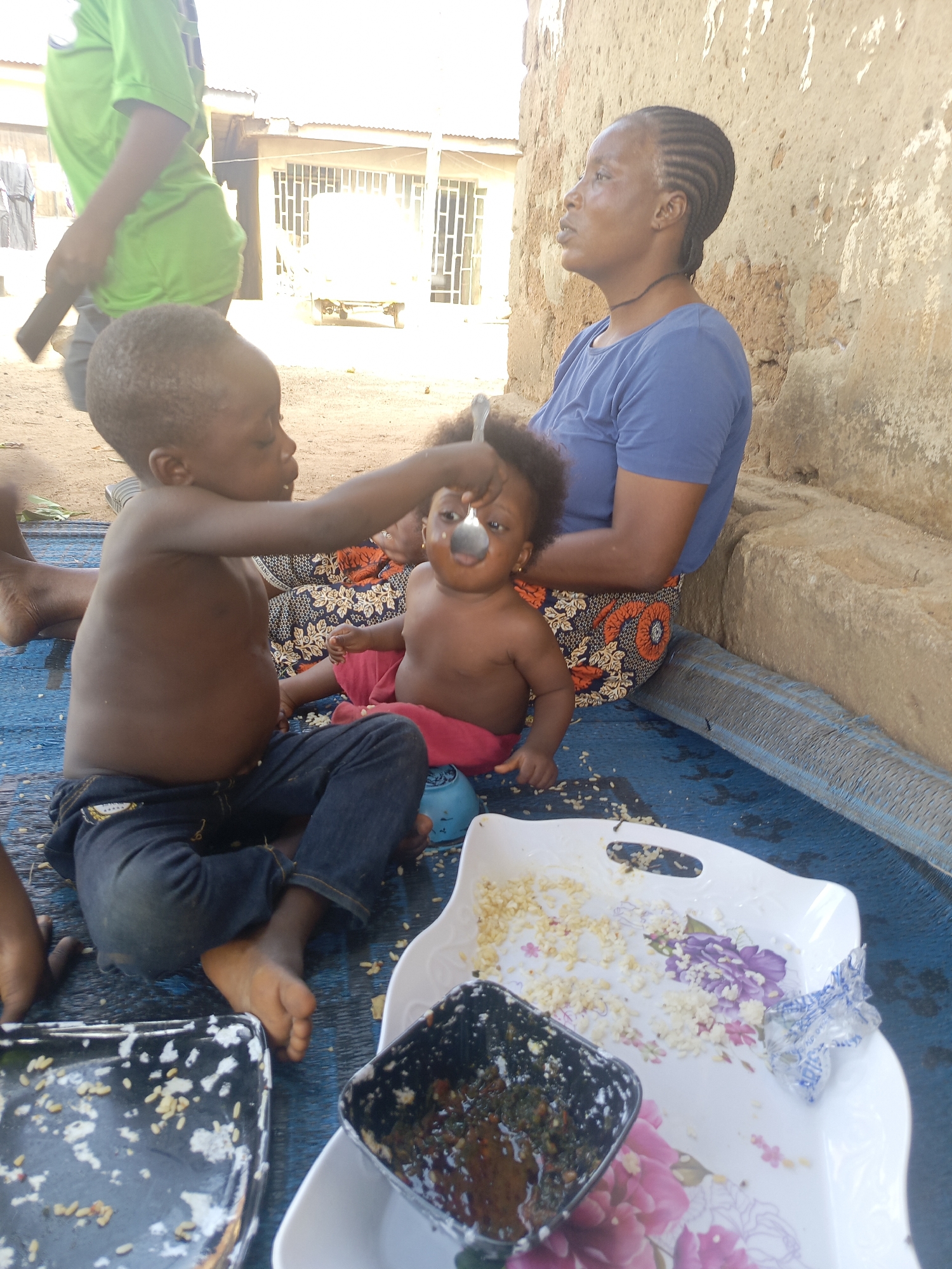 The image shows a family scene outdoors in Akwanga, Nigeria. A woman with braided hair wearing a blue shirt and patterned skirt is seated on a blue mat, next to a baby in a red skirt. A young boy wearing jeans but no shirt is sitting beside the baby and feeding her with a spoon. In front of them, on the mat, there are two trays of food; one white with floral patterns containing cooked rice, and one black containing a dark sauce. The blue mat covers the ground. To the left, a person in a green shirt is partially visible, and to the right, there is a mud or stone wall. In the background, a building with a white door and white-barred windows can be seen.