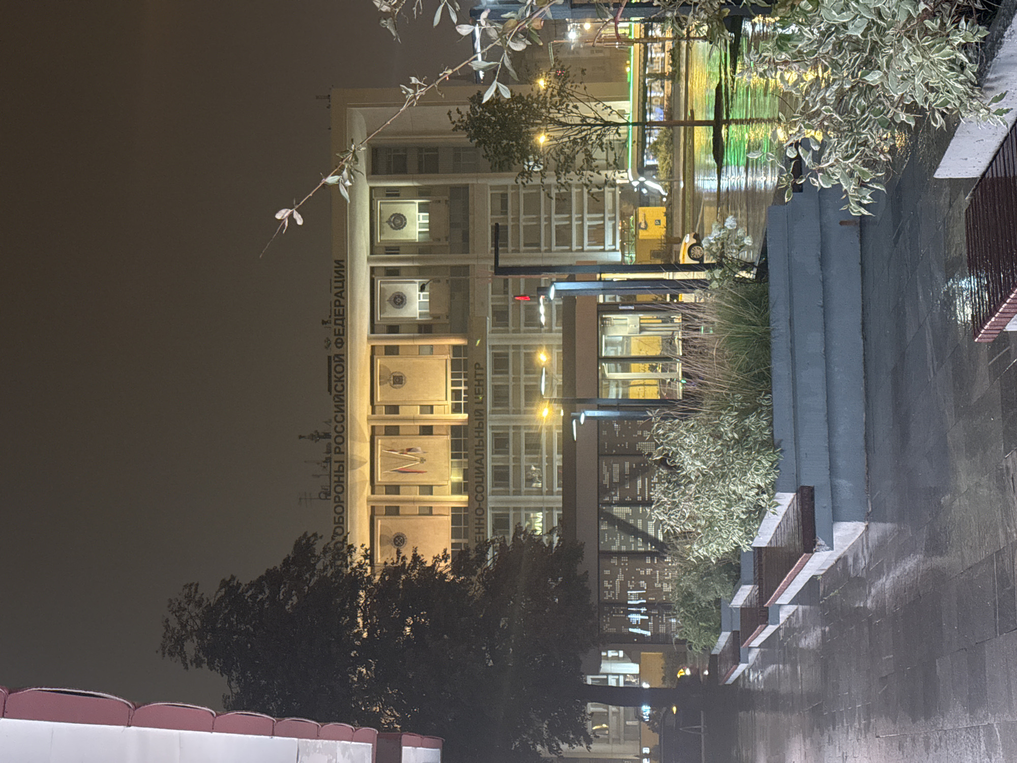 The image depicts a nighttime scene on a wet, paved street in Moscow, Russia. A large, Brutalist-style building dominates the background, illuminated by warm, yellow lights. The building features rows of windows and banners displaying emblems. Text on the building reads "ОБОРОНЫ РОССИЙСКОЙ ФЕДЕРАЦИИ" and "ЕННО-СОЦИАЛЬНЫЙ ЦЕНТР".
In the foreground, several modern concrete planters with lush green and variegated foliage are arranged along the sidewalk. Three wooden benches are placed in front of the planters, their surfaces wet from recent rain. The wet pavement reflects the ambient lights of the street.
A dark-leaved tree stands to the left of the frame, partially obscuring the building. A street lamp with a single light source illuminates the scene. In the middle ground, a yellow bus or trolley is visible, as is a white car. The overall atmosphere is one of a damp, urban evening, with the architectural features of the city illuminated against the dark sky. There are no visible people in the image.