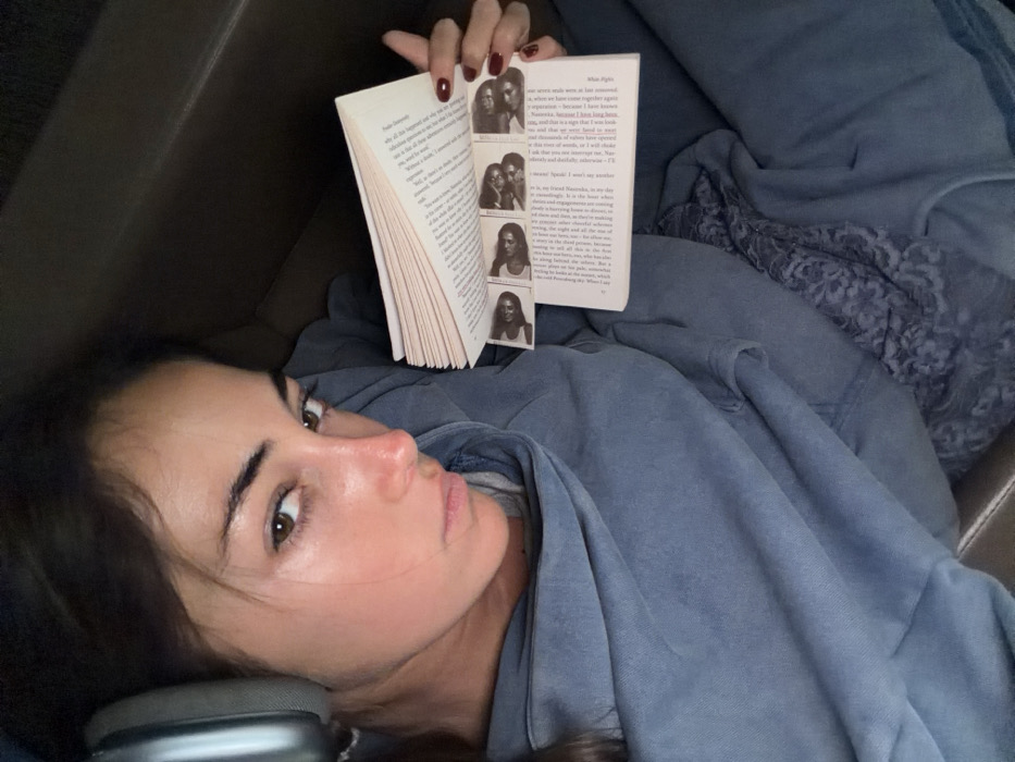 An overhead, close-up photograph captures a female subject indoors in Mexico City, Mexico. The subject, with dark hair and brown eyes, faces the camera directly with a neutral expression. She wears large, silver-grey over-ear headphones on her left ear. Her attire consists of a loose-fitting, blue-grey hooded sweatshirt and possibly matching loungewear. She holds an open paperback book in her right hand, displaying off-white pages with visible text. Inserted below the main text is a strip containing four small, horizontal black-and-white portrait photographs of women. Her right hand's fingernails are painted dark red. The background is dark and indistinct, suggesting an upholstered surface like a couch or bed.