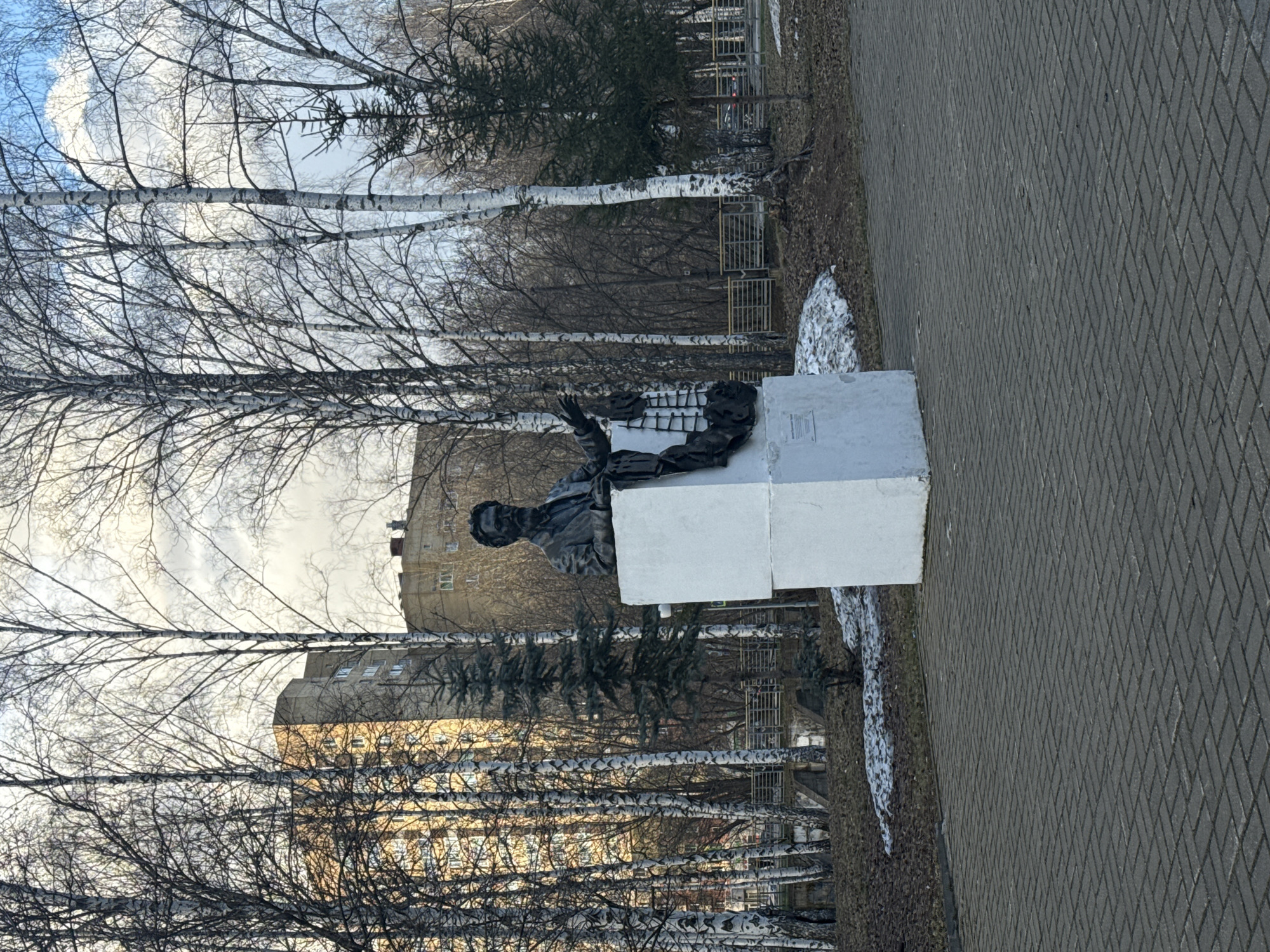 This image captures a park or public square scene in Perm, Russia, likely during late winter or early spring. The central feature is a dark-colored statue of a person, possibly male, seated on a simple white rectangular pedestal. The figure appears to be wearing formal attire and has one arm raised.
The environment is characterized by numerous bare birch trees, with their distinctive white bark and intricate network of dark, leafless branches. A few evergreen trees are also visible among the deciduous ones. The ground is a mix of bare earth and scattered patches of melting snow, reinforcing the transitional season.
A paved pathway made of grey interlocking pavers, arranged in a diamond pattern, runs alongside the statue. In the background, multi-story urban buildings are visible through the bare tree branches. One prominent building has a light-colored, possibly yellowish or beige, facade with numerous windows. Fences with a yellowish tint can also be seen in the mid-ground.
The sky is overcast but bright, indicating daylight. The scene is static, with no specific activity taking place. A small plaque, containing illegible text, is affixed to the side of the statue's pedestal, presumably identifying the monument or the person depicted. The overall impression is one of a quiet, somewhat stark urban park in a cold climate.