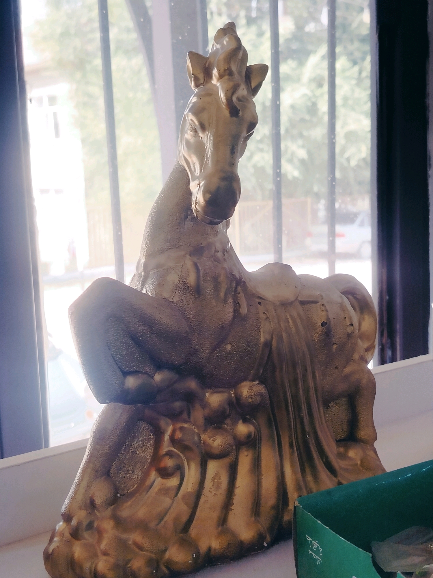 A golden statue of a horse is positioned on a white surface, likely a windowsill, in front of a window. The statue depicts the horse in a seated or kneeling posture, with its head raised. The background visible through the window shows trees and some buildings, suggesting an outdoor, possibly urban environment. The lighting indicates it is daytime, with sunlight filtering through the window, casting some glare and highlights on the statue. A corner of a green cardboard box is visible in the lower right of the frame. The location is identified as Көлкент, Kazakhstan.