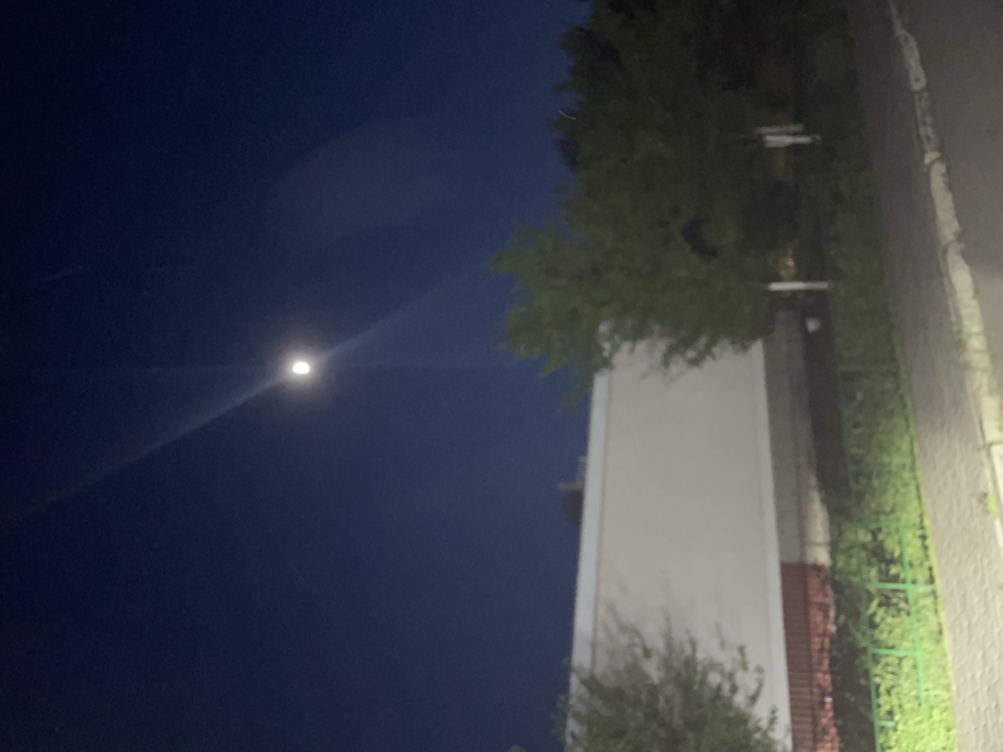 A nighttime scene in Подстёпки, Russia, showing a dark blue sky with a single bright light source, likely the moon. To the right, there's a building with a light-colored wall, partially obscured by green foliage and trees. A sidewalk or paved area runs along the right edge of the frame. No people are visible.