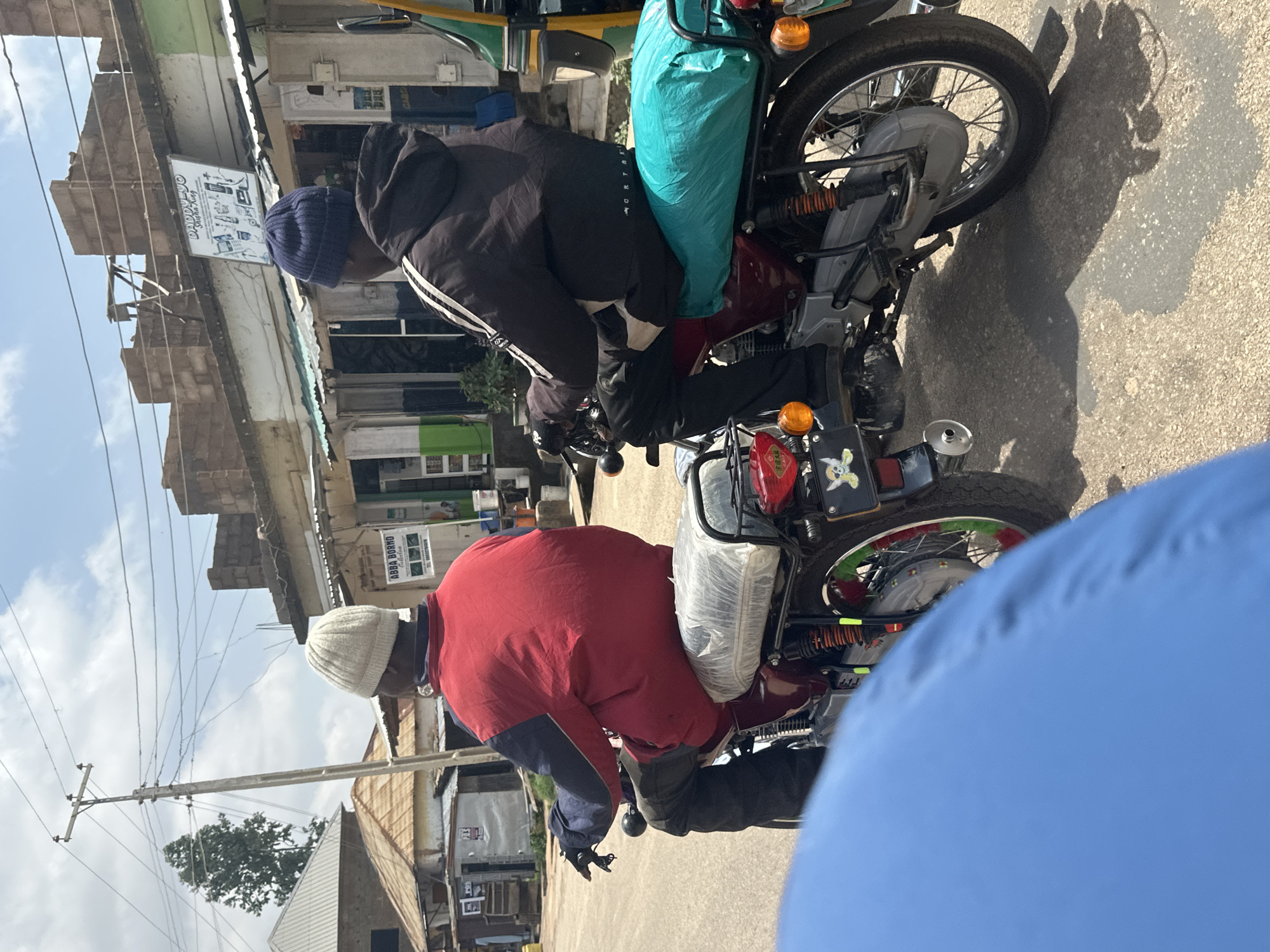 In the city of Jos, Nigeria, two individuals are seen riding motorcycles on a street. The motorcycle in the foreground has a person wearing a red and blue jacket and a white beanie. The motorcycle behind it carries a person in a black jacket with white stripes and a blue beanie, with a bright teal-colored bundle secured behind them. The background displays a row of shops with signage, including one that reads "DADDY-YO Shisha King." The street is paved, and utility poles with wires are visible against a cloudy sky. The overall scene captures a moment of daily life on a street in Jos.