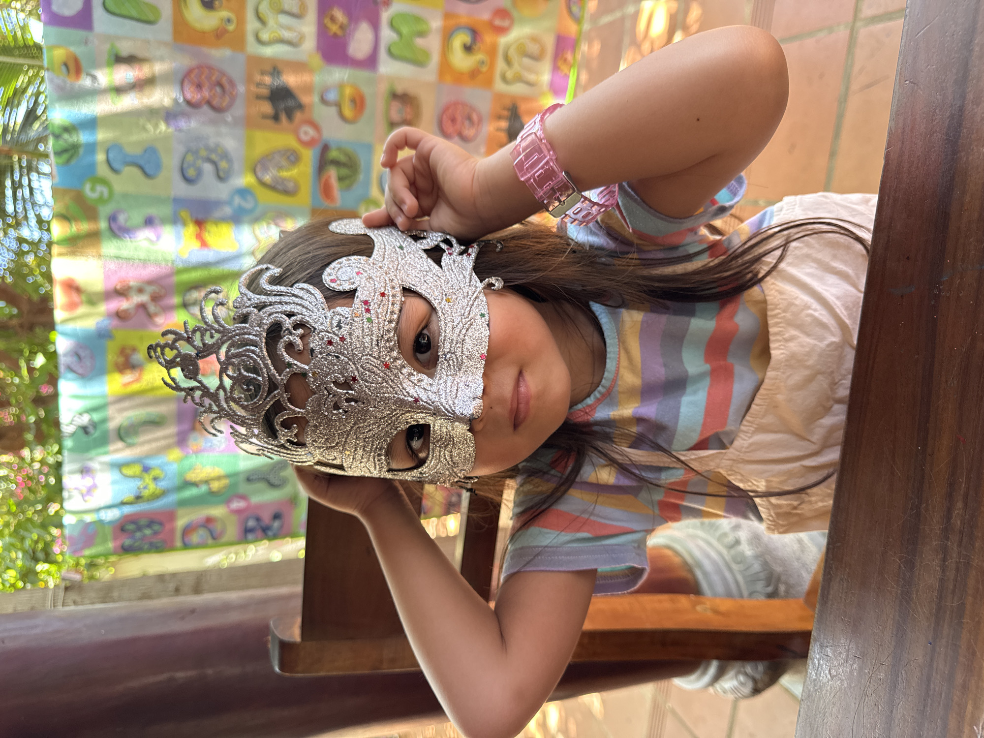 A young girl is holding a silver glitter mask up to her face. She is wearing a pink plastic bracelet and a striped shirt with a white overall dress. She is sitting at a wooden table and chair. Behind her is a colorful alphabet and picture mat. The background also includes outdoor greenery, possibly trees. The location may be Mũi Né, Vietnam.