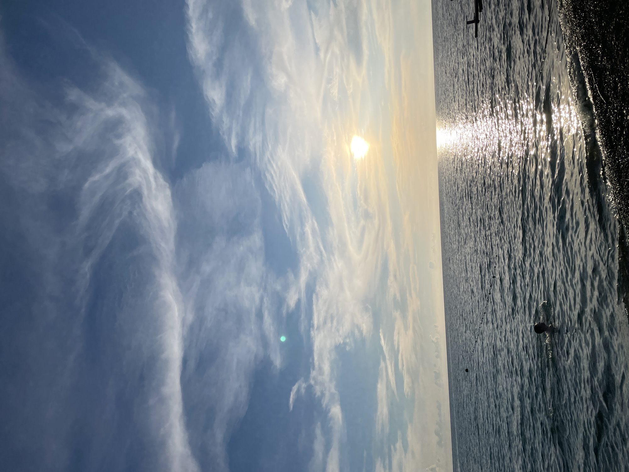 The image depicts a serene view of the Black Sea at sunset, likely near Sochi, Russia. The sky is a canvas of blue with wispy cirrus clouds painted across it, catching the golden hues of the setting sun. The sun itself is a bright orb, casting a shimmering reflection on the water's surface. The sea is calm, with gentle waves lapping at a pebbly shore in the foreground. In the water, a person is swimming, their head and shoulders visible above the surface. Further out in the sea, a line of buoys marks a designated swimming area. A small portion of a wooden structure or pier is visible on the right side of the frame, suggesting a recreational area. The overall atmosphere is peaceful and tranquil, capturing the essence of a summer evening by the sea.