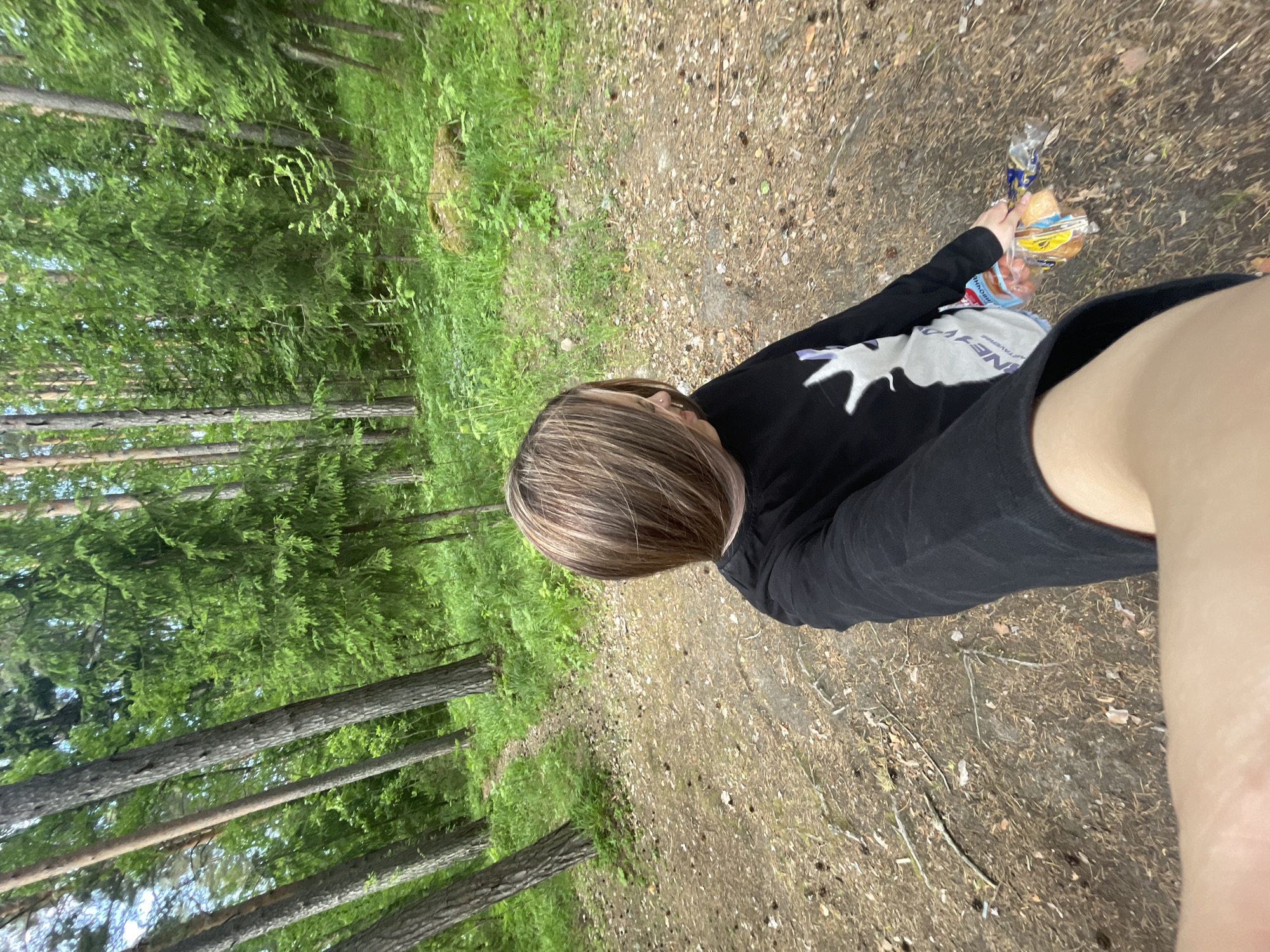 The image is a first-person perspective selfie captured in a dense, lush green forest environment, specifically located within or adjacent to Boitsovo, Russia. The foreground and midground feature a narrow, unpaved dirt path covered with pine needles, small fallen branches, and occasional patches of sparse green grass. The path recedes into the background on the left, flanked by numerous tall, slender coniferous trees with textured bark. The surrounding forest is rich with vibrant green foliage, including younger evergreen trees and dense undergrowth, particularly on the gently sloping terrain to the right. A large, moss-covered boulder is visible among the greenery on the mid-right side of the frame.
The subject is an individual with medium-long, straight brown hair featuring blonde highlights, styled with a side part that partially obscures the face. A silver piercing is visible on the left nostril. The person is wearing a long-sleeved black shirt with a large white graphic on the front, depicting a silhouette, possibly an animal head. Their right arm extends into the foreground, holding a transparent plastic bag containing various individually wrapped food items, including multiple orange-colored sausages in clear packaging and several baked goods or buns. Cyrillic text, notably "ПРОДУКЦИЯ" (Produktsiya - Products), is visible on the sausage packaging. A light-colored reusable tote bag with dark text, including "НЕХВАТ TAAVERNA," is slung over the subject's left shoulder. The individual is looking slightly downward and to their right, seemingly at the items they are holding or the immediate path ahead.