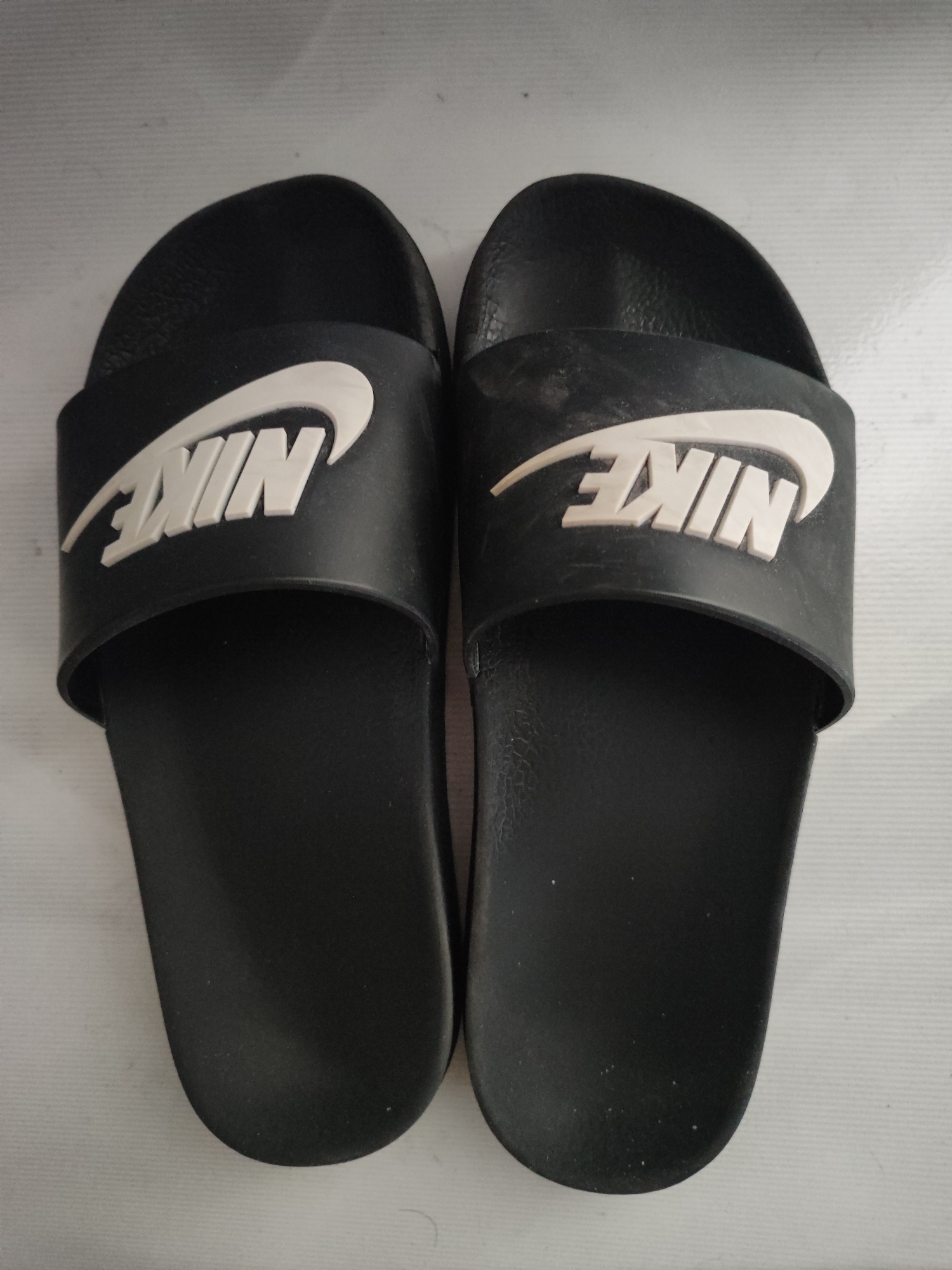 This is a top-down, eye-level, full-shot, medium-full photograph of a pair of black Nike slide sandals. The sandals are placed side-by-side on a white, textured surface. Each sandal features a broad, black strap with a white, raised Nike logo. The lighting is even and diffused, casting no significant shadows. The background is a plain white surface with a subtle ribbed texture. The image's quality is clear, with no visible dust or debris on the sandals. The scene is static and unpopulated, suggesting an indoor setting. The location of Snezhinsk, Russia, is not visually represented in the image; this information is external to the visual content.