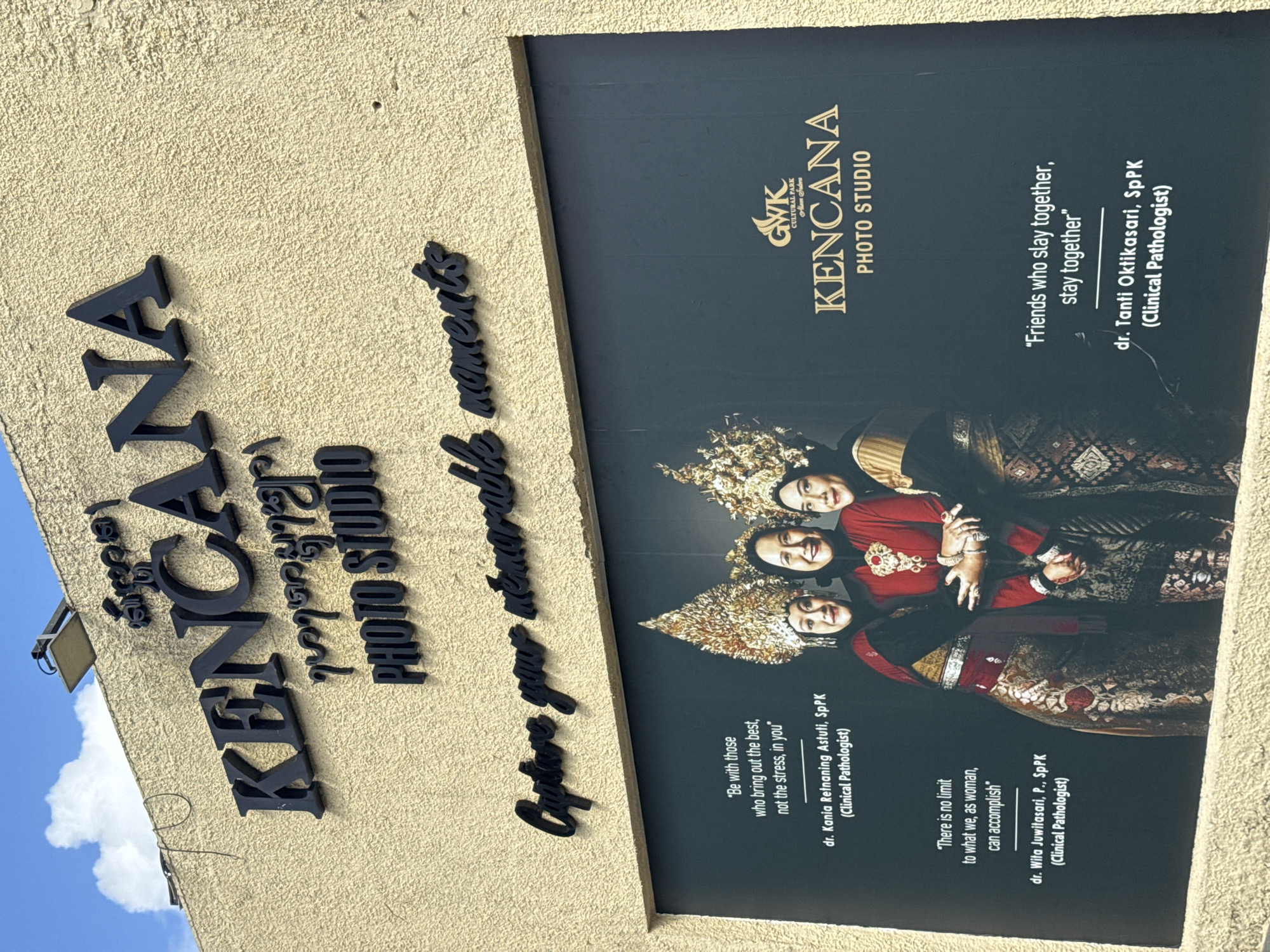 The image shows a building facade displaying the name of a photo studio, "Kencana," in Kuta Selatan, Indonesia. The studio's name appears both directly on the building in black lettering and on a large black sign, which also includes the phrase "Capture your memorable moments." The sign features an image of three women dressed in traditional Indonesian attire, likely related to weddings or other formal occasions. The sign also includes three quotes about empowering women and friendships, each credited to a different female doctor with the qualification "Clinical Pathologist." The sign also features the GWK Cultural Park logo and a tagline "Alam Srders."