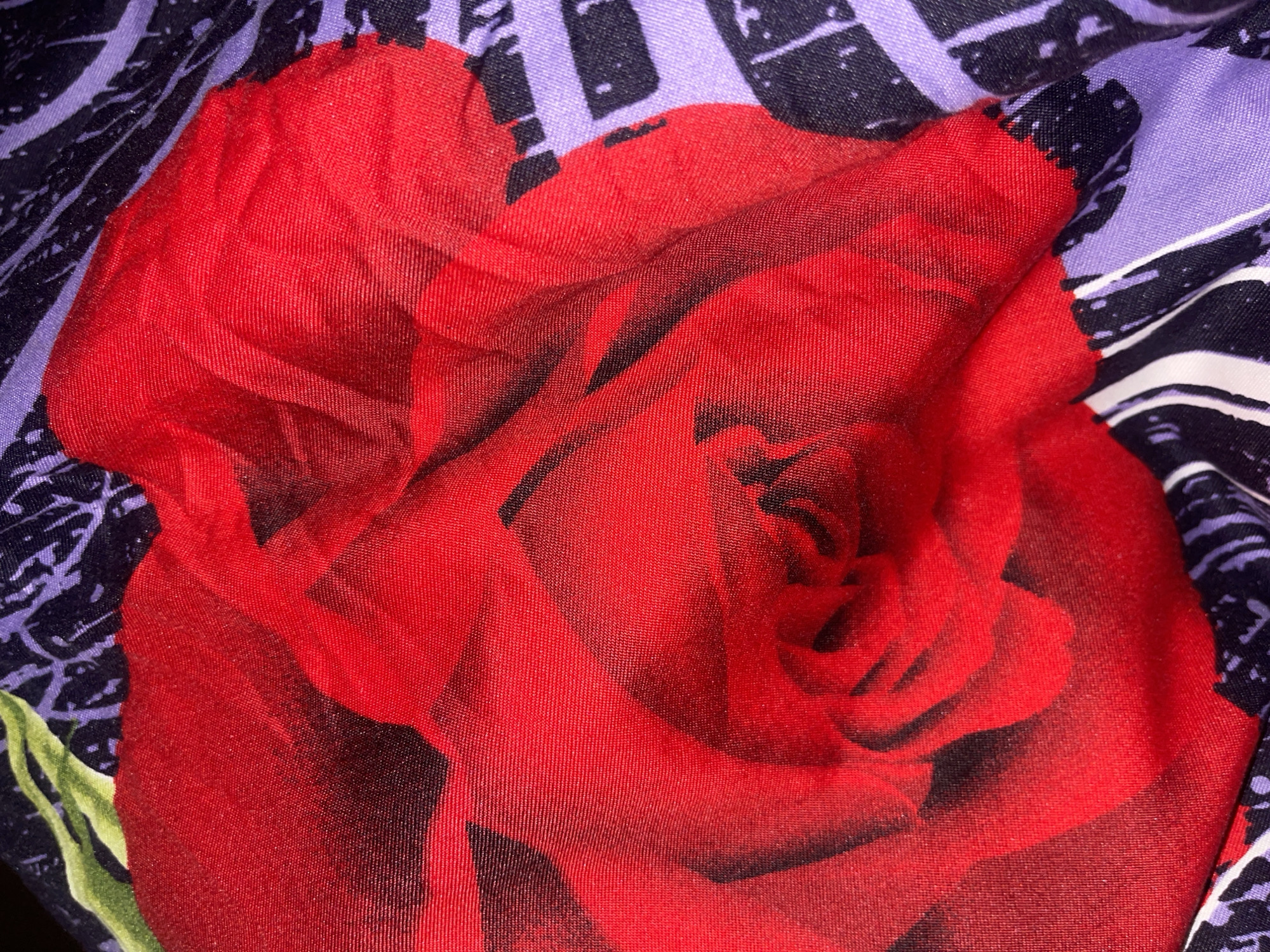 The image is a close-up, overhead shot of a red rose printed on fabric, with a purple and black abstract pattern in the background. The fabric has a slight sheen and is creased in places, creating shadows and highlights on the rose petals. The rose appears to be a large, stylized design rather than a realistic depiction. There are no people or discernible actions present in the image. The scene is isolated to the fabric itself. No information about its location in Minsk, Belarus is available from the visual content.