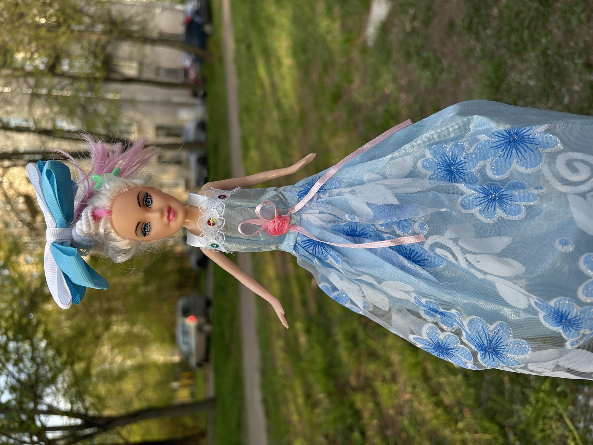 A close-up, outdoor, full shot of a doll is seen lying on a green lawn. The doll has blonde hair with pink streaks, a large blue and white bow in its hair, and blue eyes. It wears a light blue dress with white and blue flower patterns, a white lace collar, and a pink ribbon belt with a small pink flower. The lawn is green with trees in the background. A sidewalk and blurred building are also visible. The location appears to be a park or grassy area in a city.
While it is impossible to confirm the exact location from the image, nothing in this image suggests it is in Saint Petersburg, Russia.