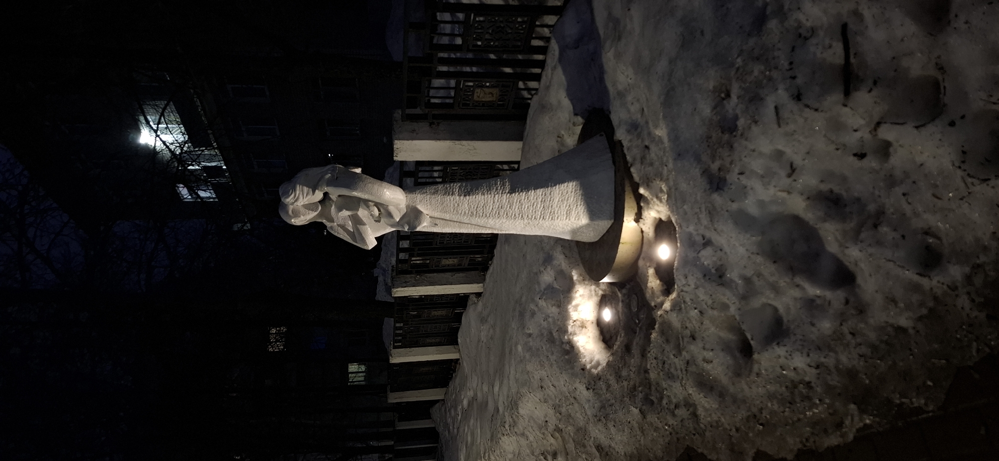 The image captures a sculpture in Chaikovsky, Russia, at night. The sculpture, a white abstract figure, is situated outside, near a staircase with ornate metalwork. The sculpture is embedded in a snowdrift, illuminated by two light sources from within the snow. The surroundings are dark, with the outlines of buildings and trees visible in the background.