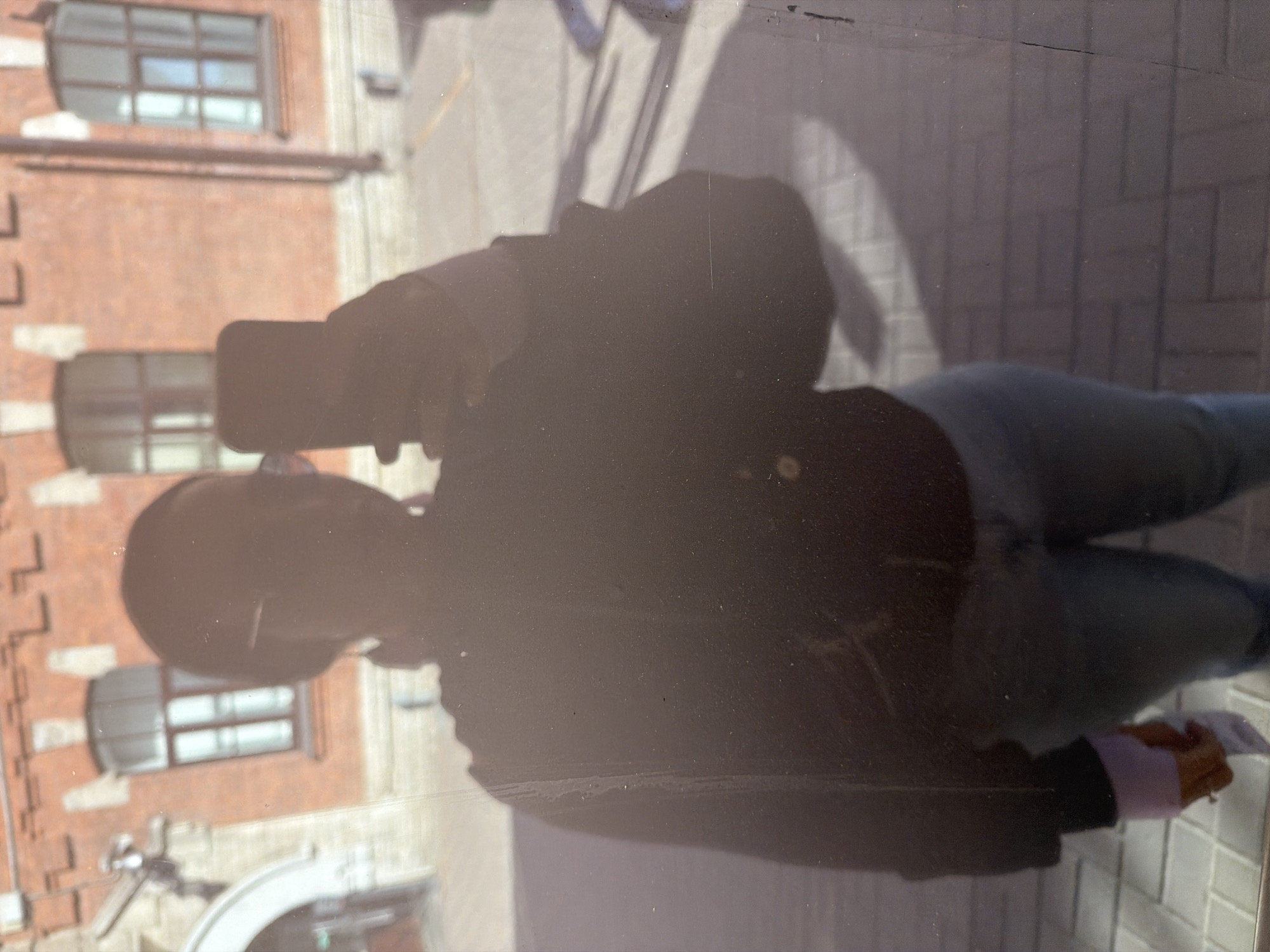 The image depicts a person reflected in a window while holding a phone to take a picture. The reflection shows a red brick building with arched windows, a brick-paved sidewalk, and the person wearing jeans and a dark jacket. The location is likely Saint Petersburg, Russia, based on the architectural style of the building, especially the windows and the brick facade. The person is standing outside and using their phone to capture the scene.