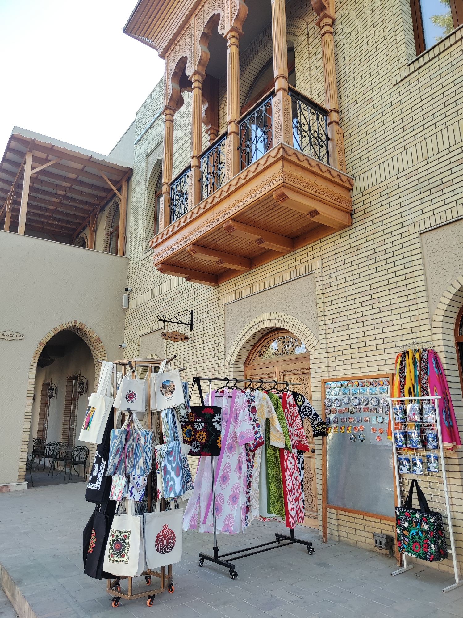 In front of a tan brick building with traditional Uzbek architectural elements, including a carved wooden balcony supported by columns and an arched doorway, a street vendor displays a variety of goods. The building features intricate wooden carvings and arched windows. In the foreground, a rack of colorful textile products, primarily patterned tote bags and flowing garments, is set up for sale. A separate display board exhibits a collection of small decorative plates and magnets. The ground is paved with interlocking stones. The scene is outdoors with daylight suggesting it is daytime. The overall setting is likely a marketplace or a street in Tashkent, Uzbekistan, given the architectural style.